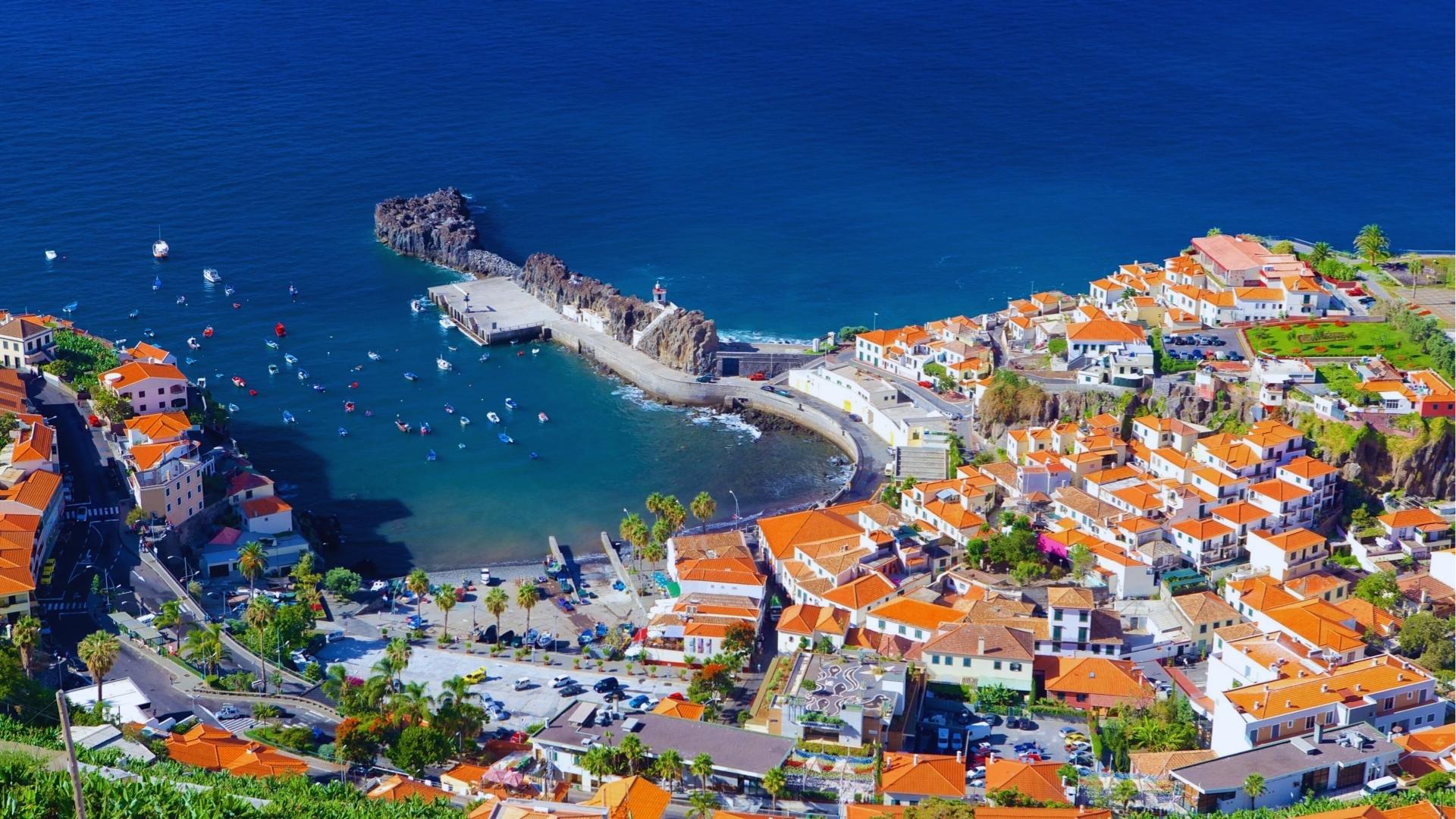 Aerial view of Câmara de Lobos fishing village in Madeira with terracotta rooftops, harbor pier, boats, and deep blue Atlantic.