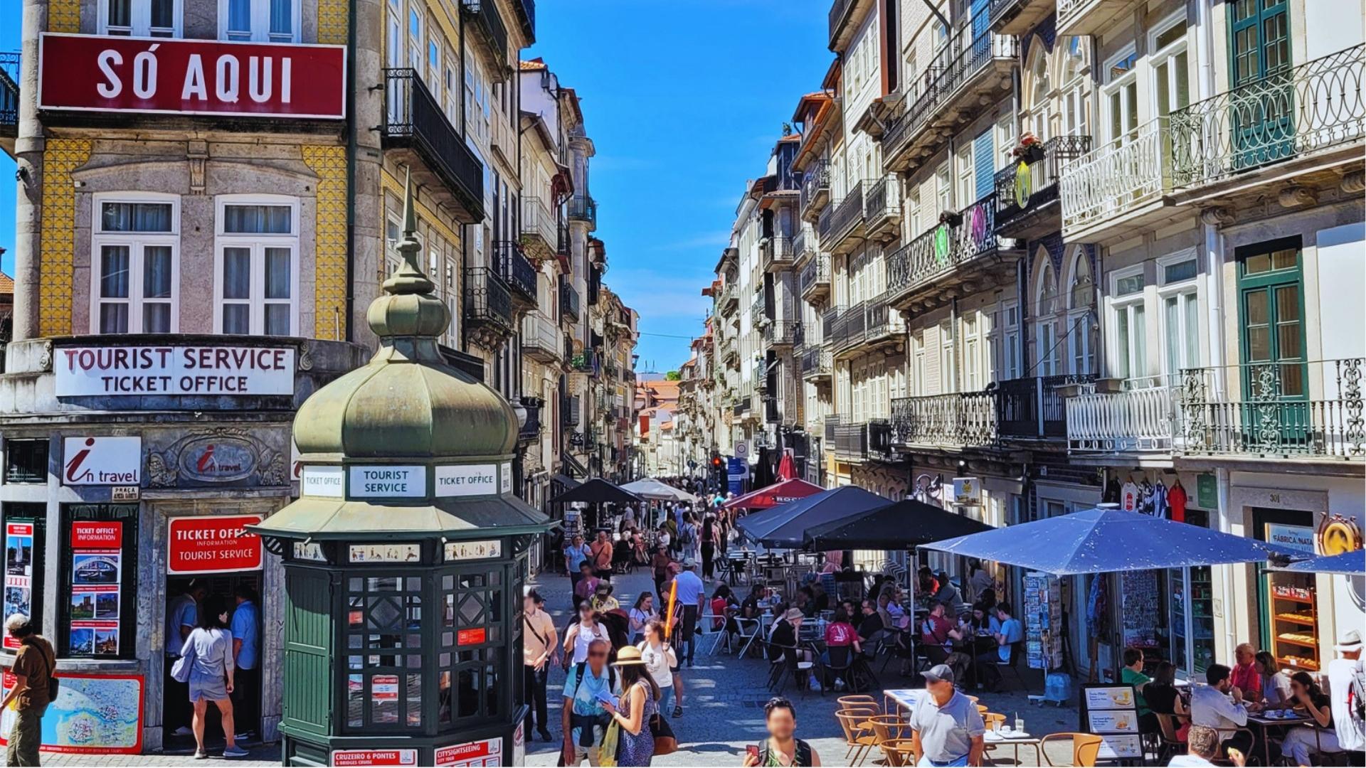 Bustling Rua das Flores in Porto with outdoor cafes, ticket office kiosks, colorful tiled buildings, and tourists on sunny days.