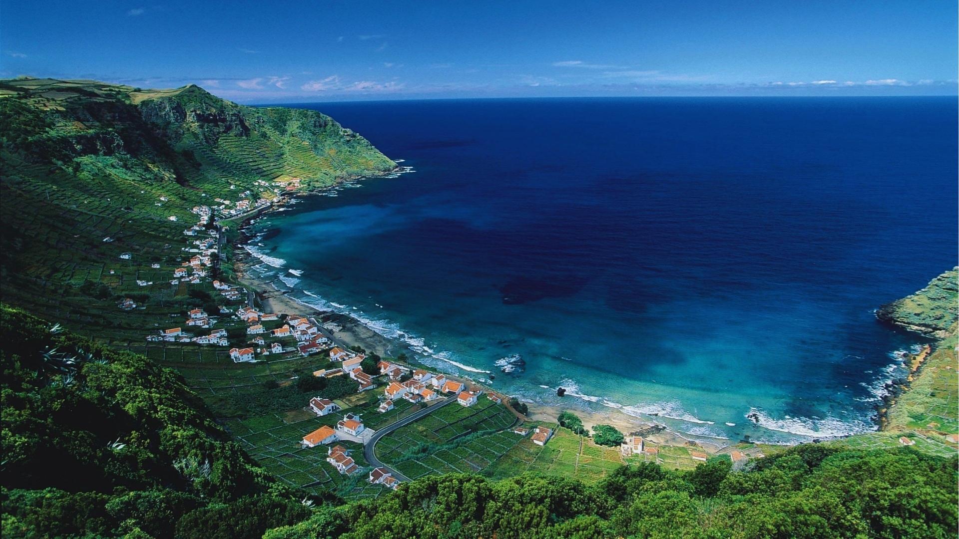 Coastal village in the Azores with white houses, red roofs, terraced vineyards, and curved sandy beach against lush green cliffs.