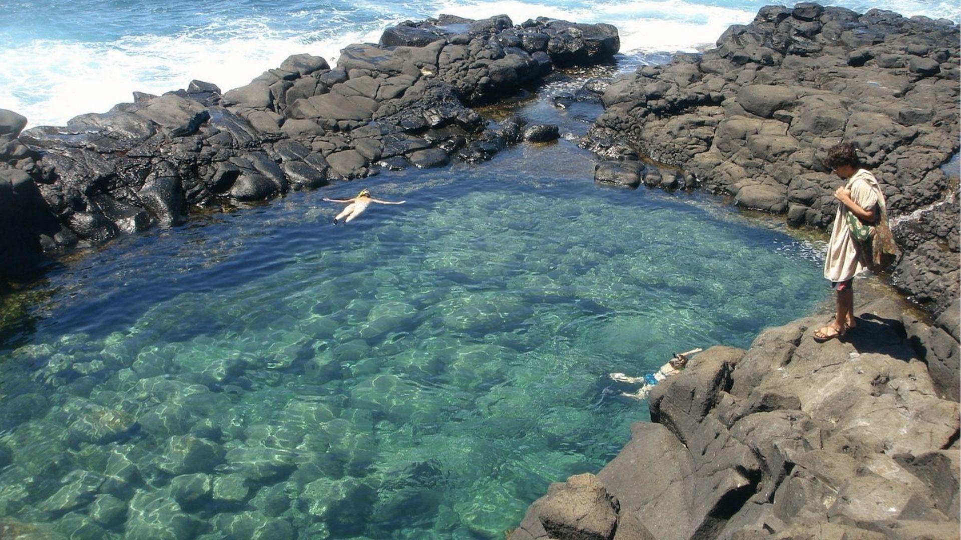 Swimmers float in a clear tide pool at Queen's Bath, reached by a short cliff trail on Kauai.