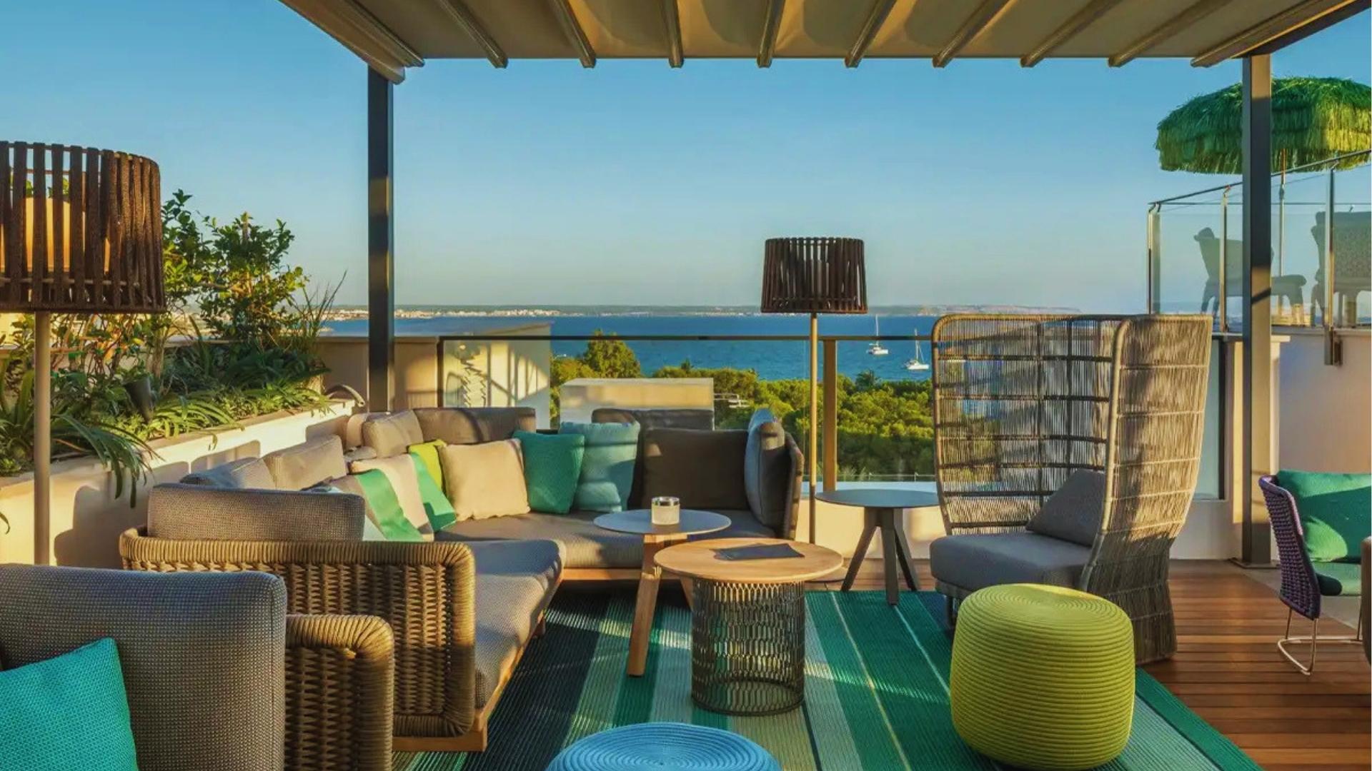 Modern rooftop lounge with wicker chairs, striped rug, and ocean view of Palma Bay with sailboats under blue sky.