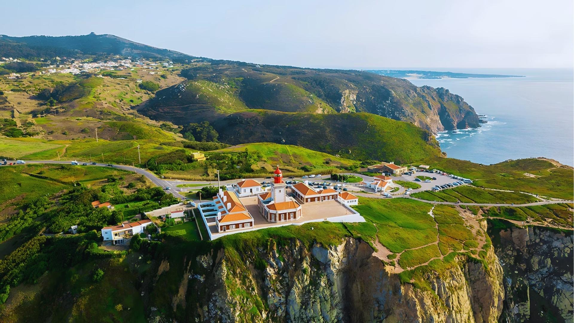 Dramatic aerial of Cabo da Roca lighthouse perched on green cliffs above Atlantic Ocean at Portugal’s westernmost point.