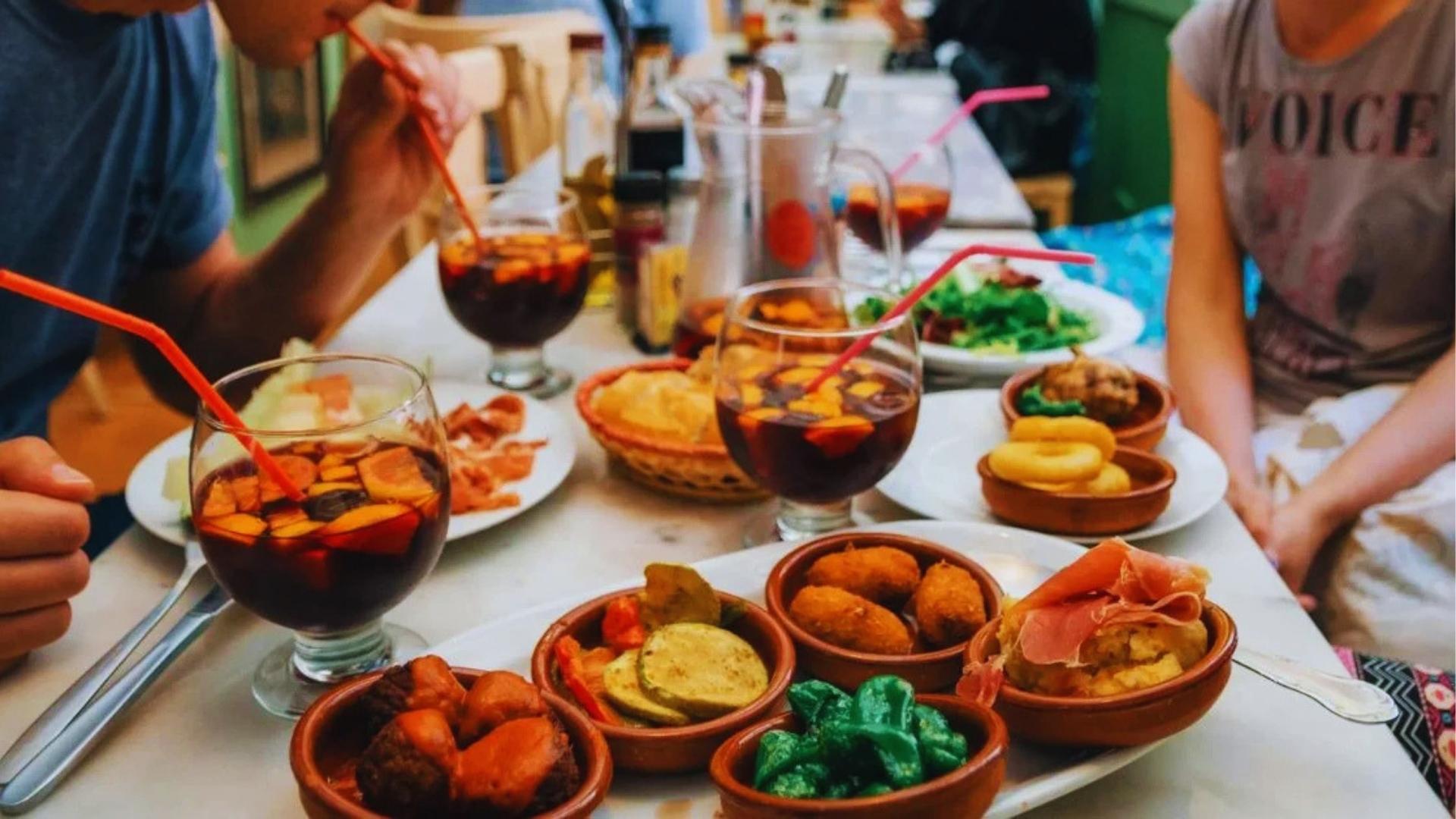 Table filled with Spanish tapas, sangria glasses with orange slices, croquettes, padron peppers, and jamón at a lively restaurant.