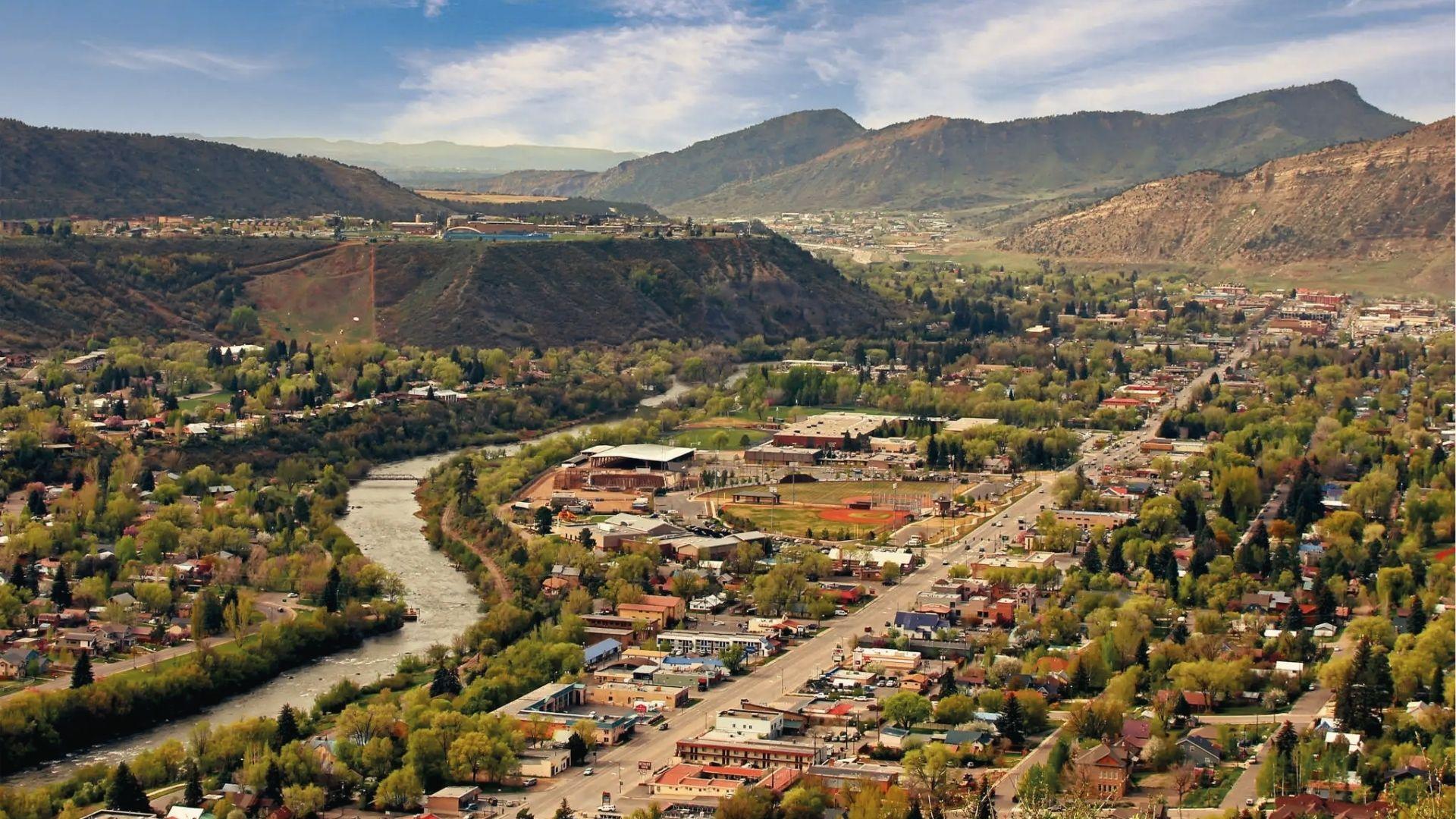 River winds through a green valley town framed by mountains. Cozy December gateway to snow activities.