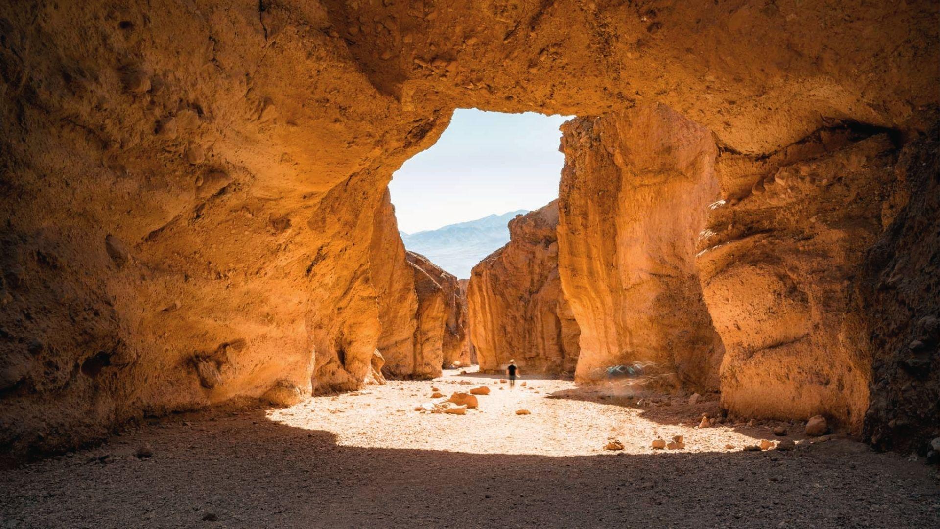 Hiker walks through sunlit canyon walls as spring light glows in Death Valley’s Golden Canyon.