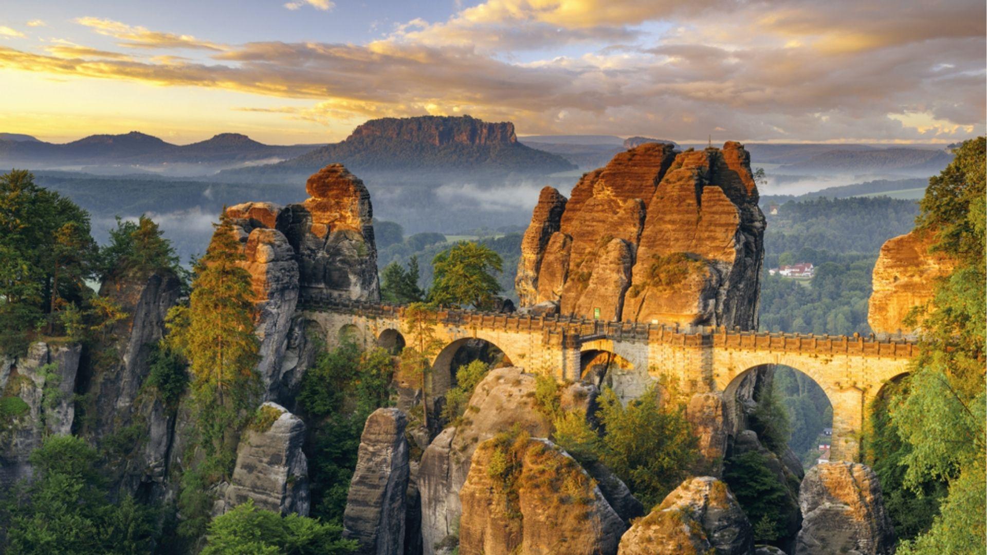 Stone bridge arches across sandstone cliffs at sunrise, misty valleys reveal Saxony’s secret spot.