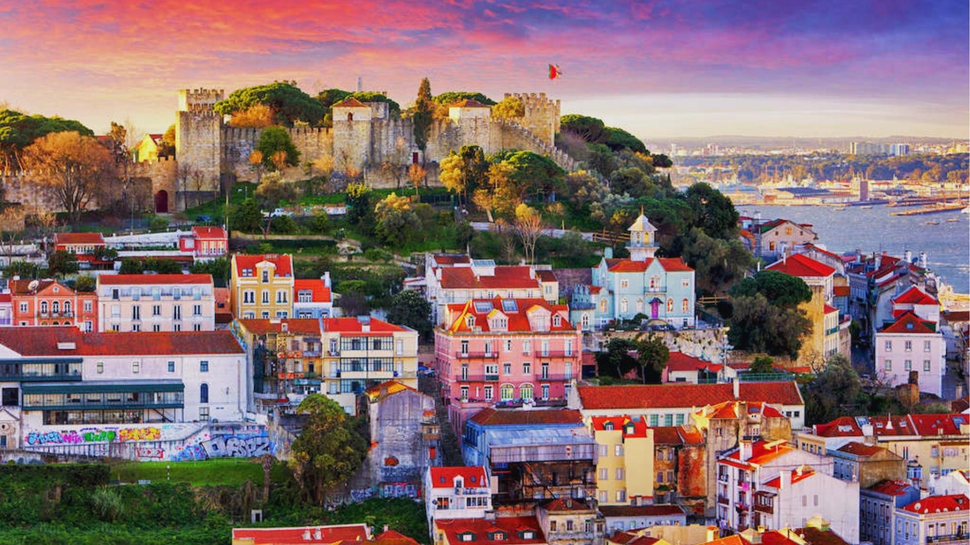 Sunset over São Jorge Castle hilltop with pastel Lisbon buildings, red rooftops, and Tagus River under pink purple sky.
