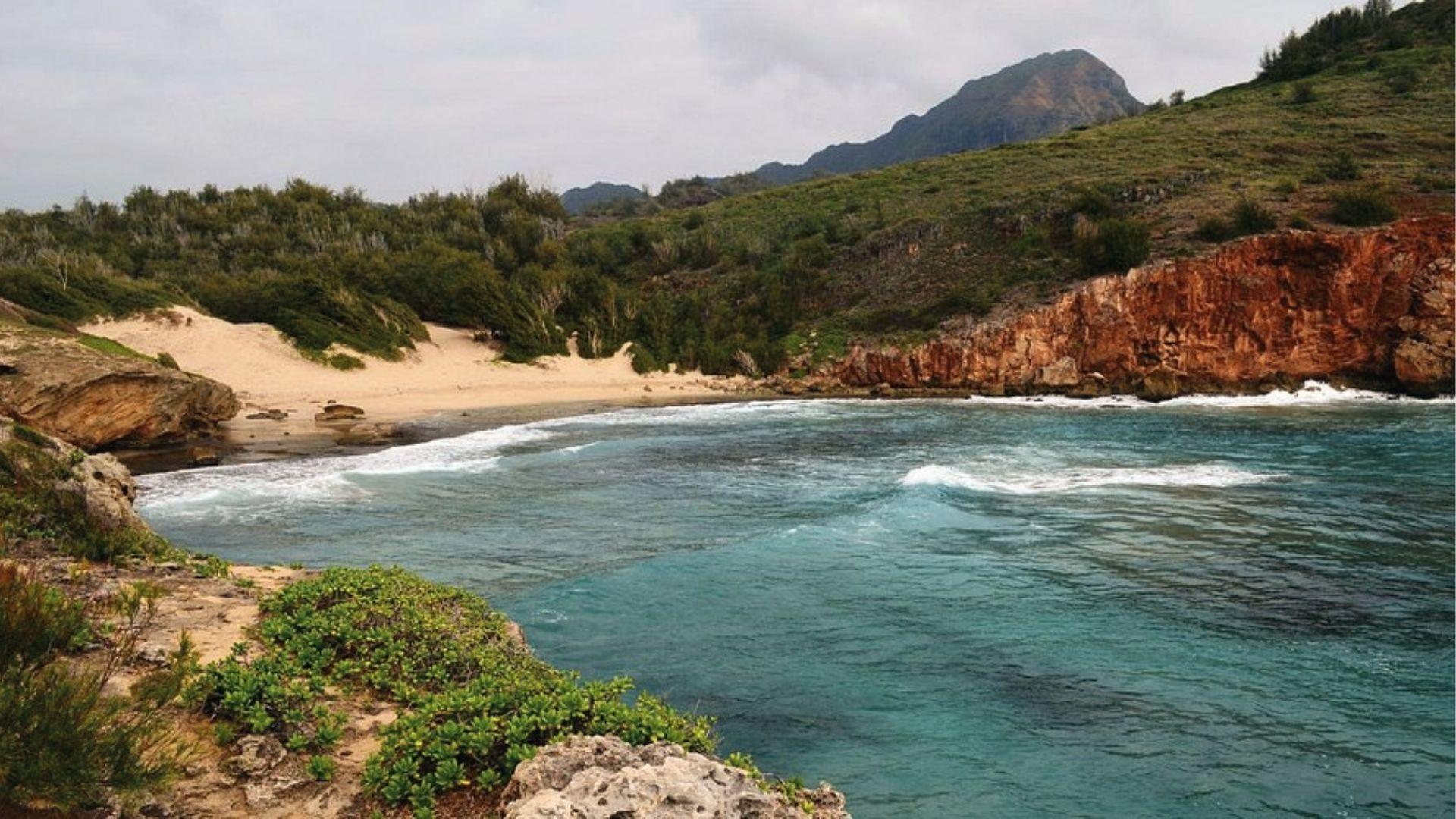 Secluded Mahaulepu Beach with red cliffs and waves, end of coastal hike in south Kauai.