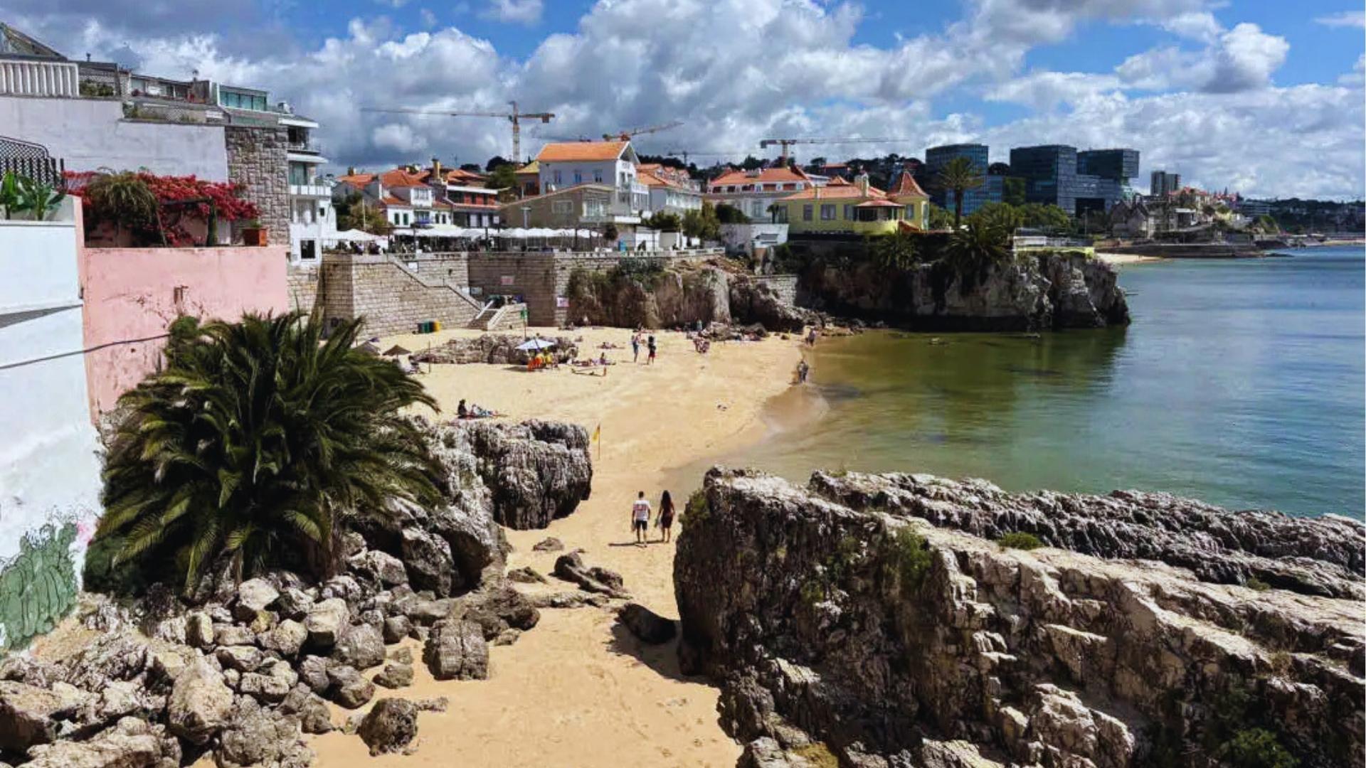Small sandy beach cove in Cascais with rocky cliffs, palm trees, and pastel buildings under blue sky with scattered clouds.