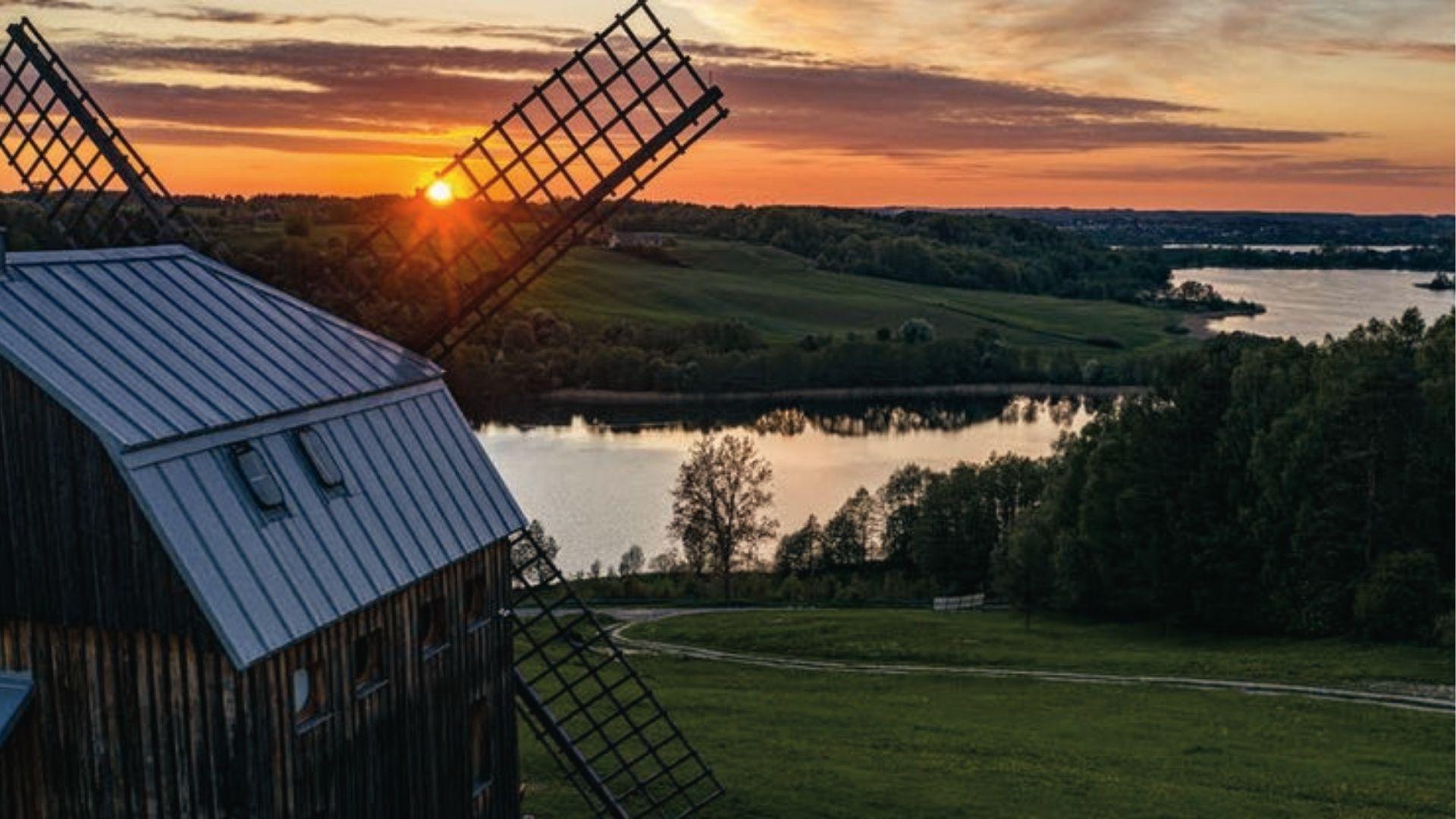 Windmill blades catch golden sunset over calm lake, Lithuania’s countryside reveals quiet, pastoral charm.