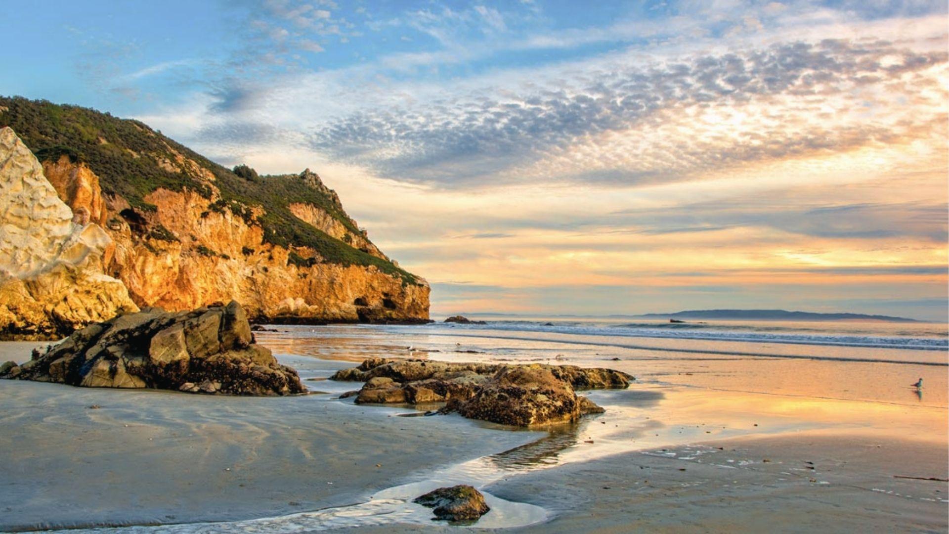 Golden sunset lights up rocky cliffs and calm waves on shore. Peaceful December beach escape.