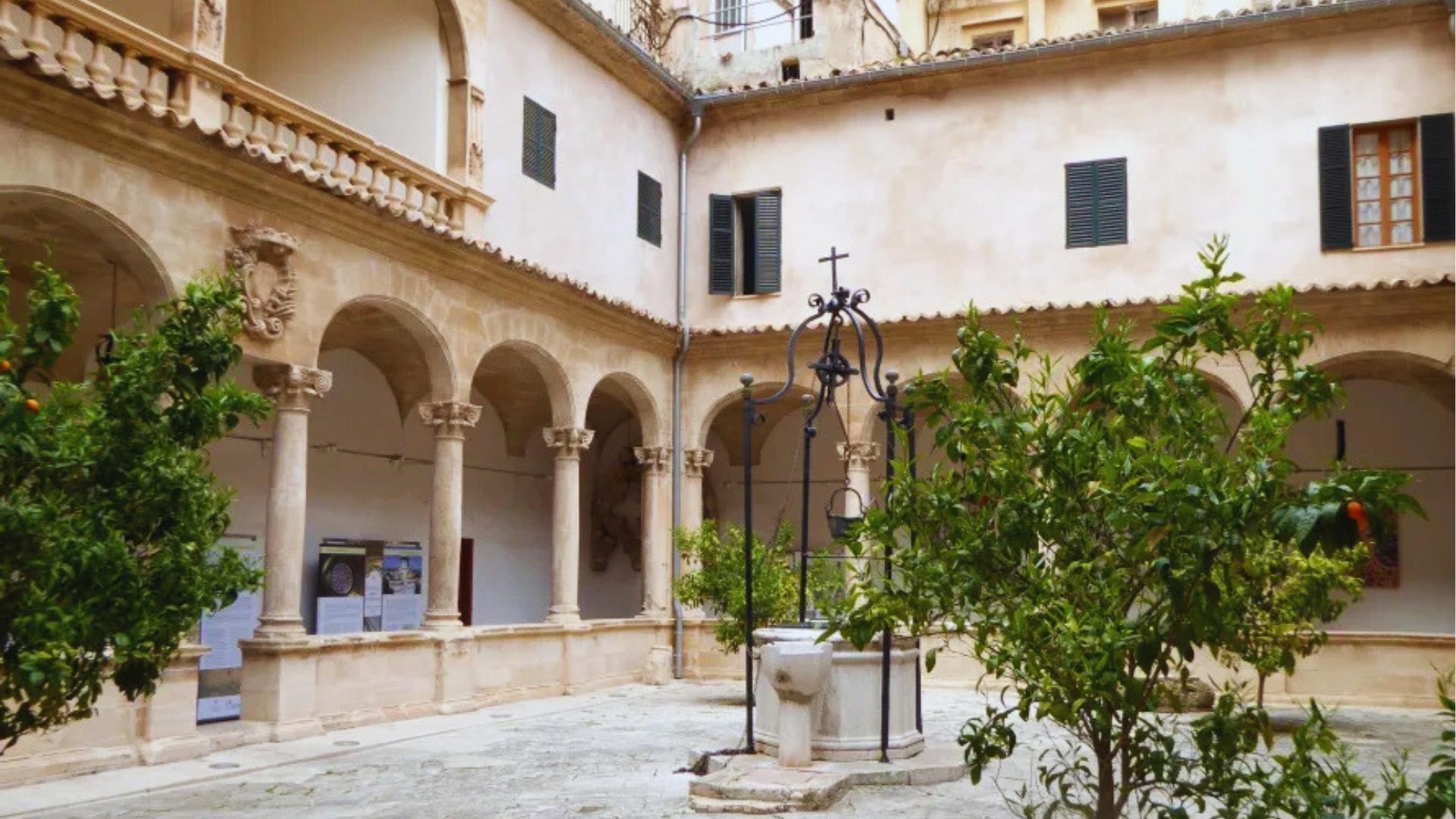 Peaceful stone courtyard with arched colonnades, iron well, and orange trees at historic monastery in Mallorca, Spain.