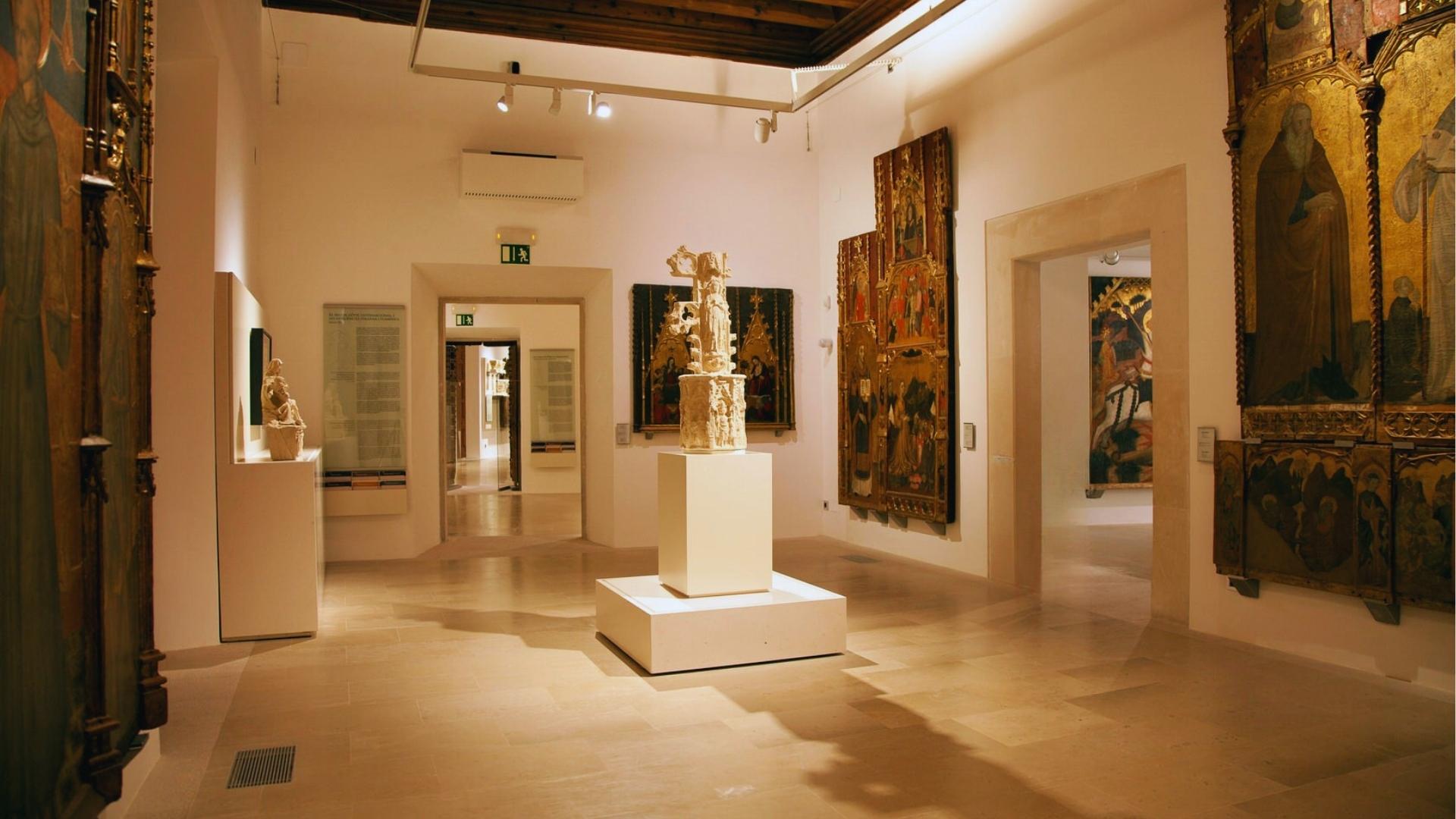 Museum gallery displaying medieval religious altarpieces and stone sculpture on white pedestal with warm lighting and tiled floors.