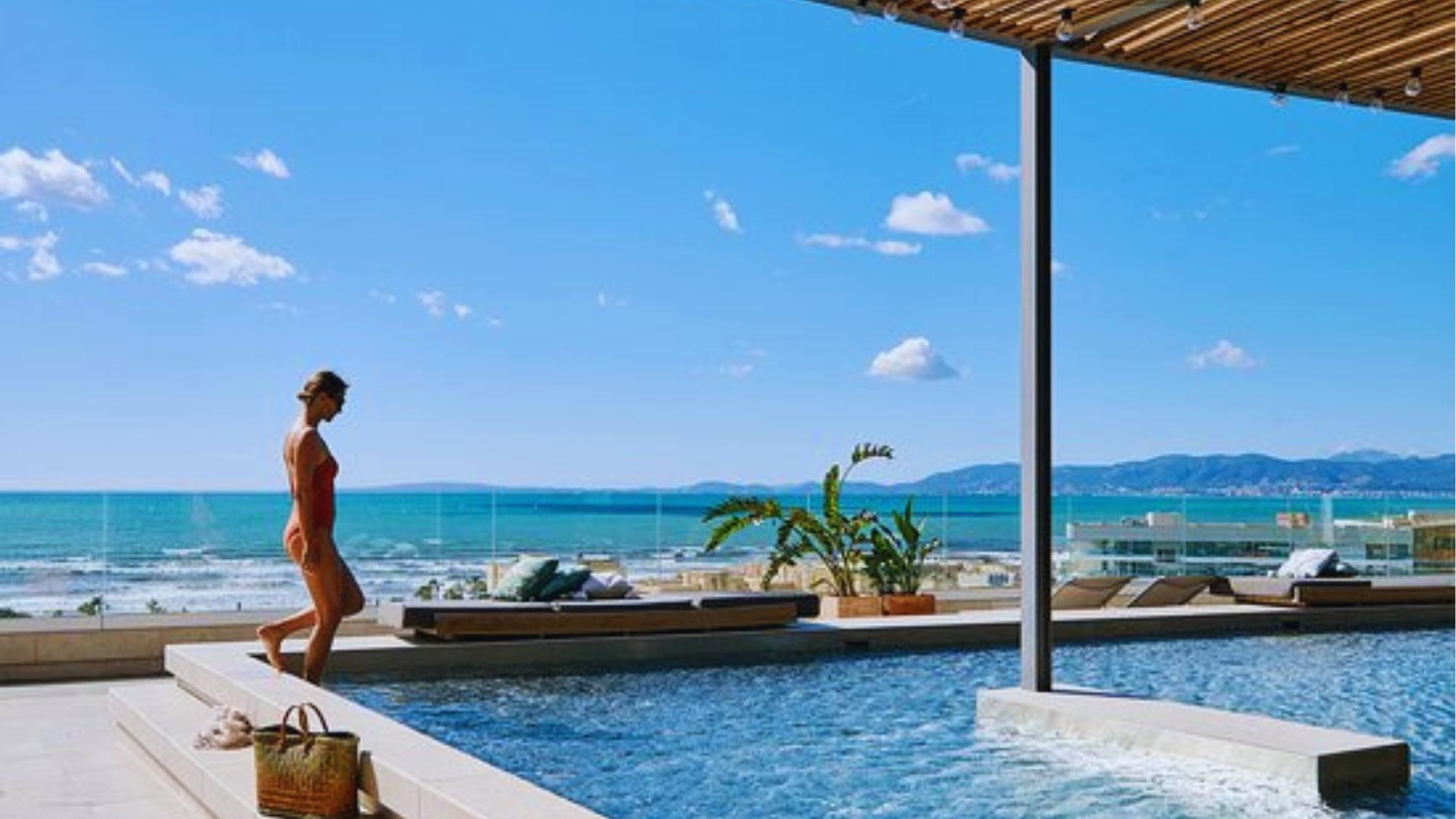 Woman in red swimsuit steps into rooftop infinity pool overlooking turquoise ocean and mountains on sunny Mallorca day.