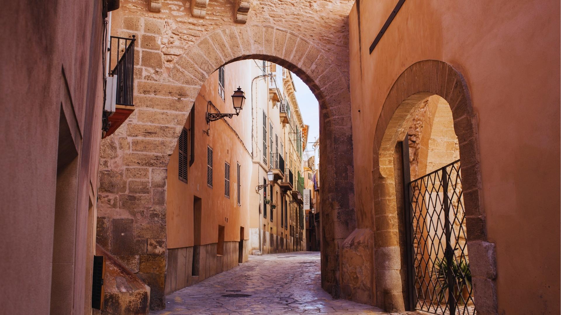 Narrow cobblestone alley in Palma Old Town framed by stone arch, terracotta walls, wrought-iron balconies, and vintage street lamps.