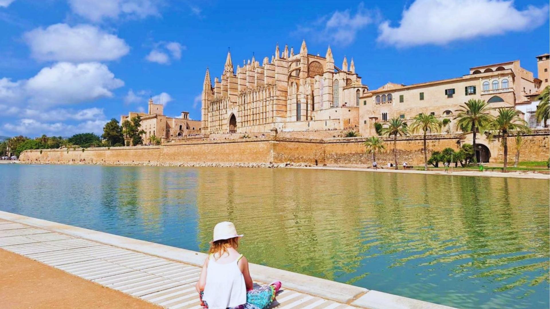 Child in sunhat sits by Parc de la Mar lake gazing at Palma Cathedral and Royal Palace under bright blue sky.