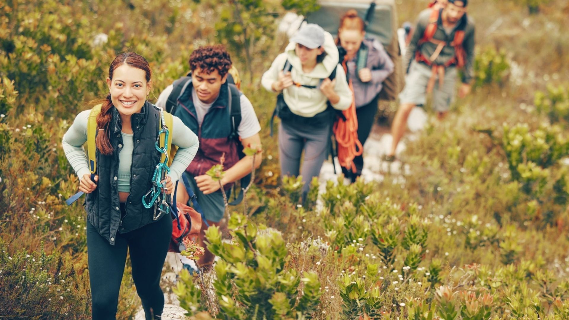 Group of smiling hikers with backpacks ascends through wildflowers and shrubs on summer trail near Ice Lake Colorado