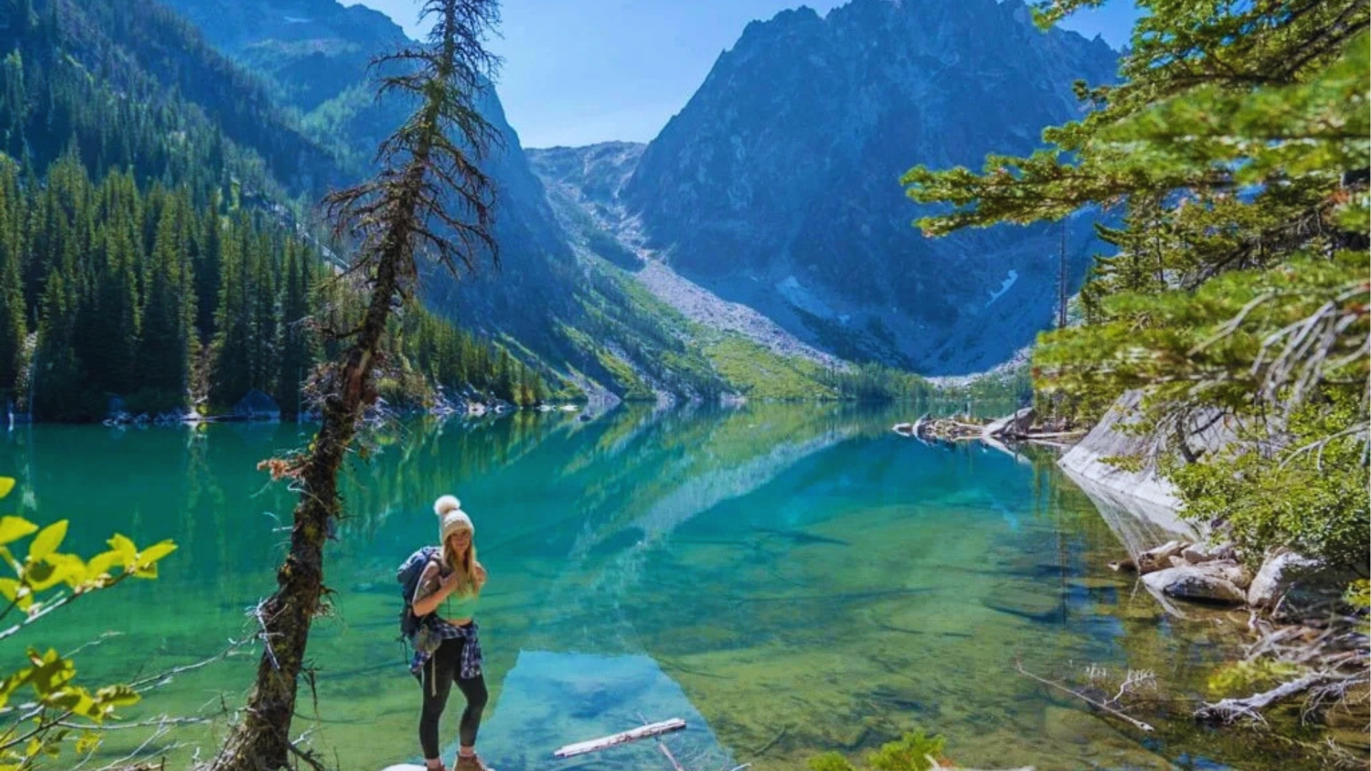 Hiker with white beanie and backpack stands on lakeshore as calm Colchuck Lake reflects pine trees and Dragontail Peak.