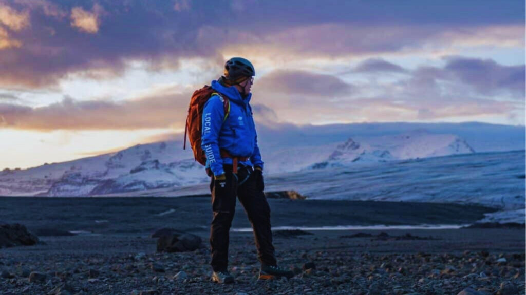 Local guide in blue jacket and helmet stands on black volcanic rock with orange backpack overlooking Iceland glacier at sunset.