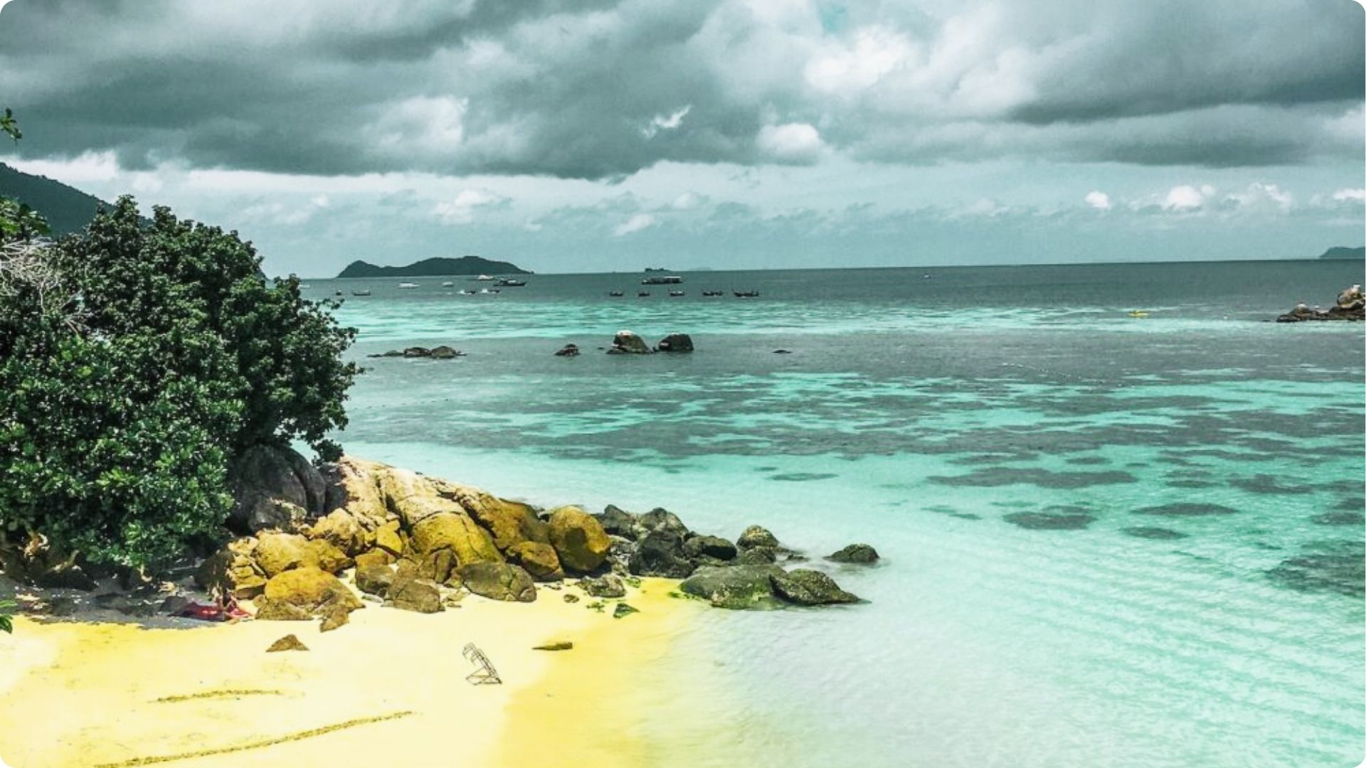 Yellow sand and large boulders line a quiet Thai beach with shallow turquoise water.