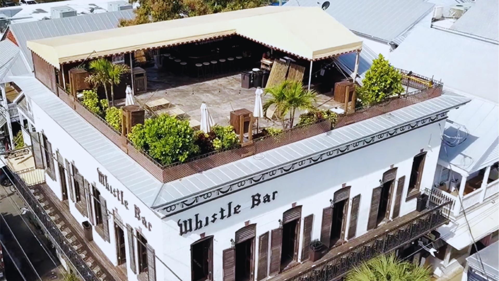 Aerial view of Whistle Bar rooftop deck with plants, umbrellas, and canopy on historic white Key West building.