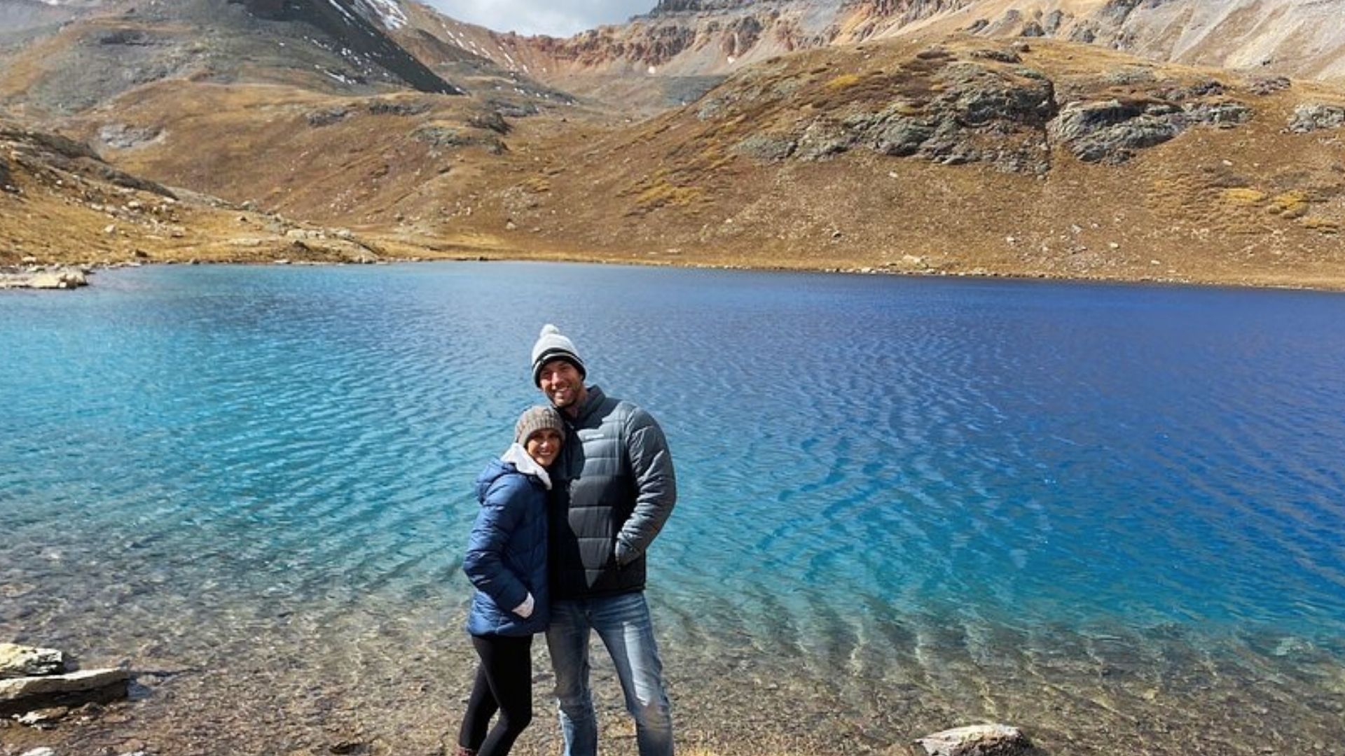 Smiling couple in winter jackets stands at Ice Lake Colorado shoreline with bright blue water and brown mountain backdrop.