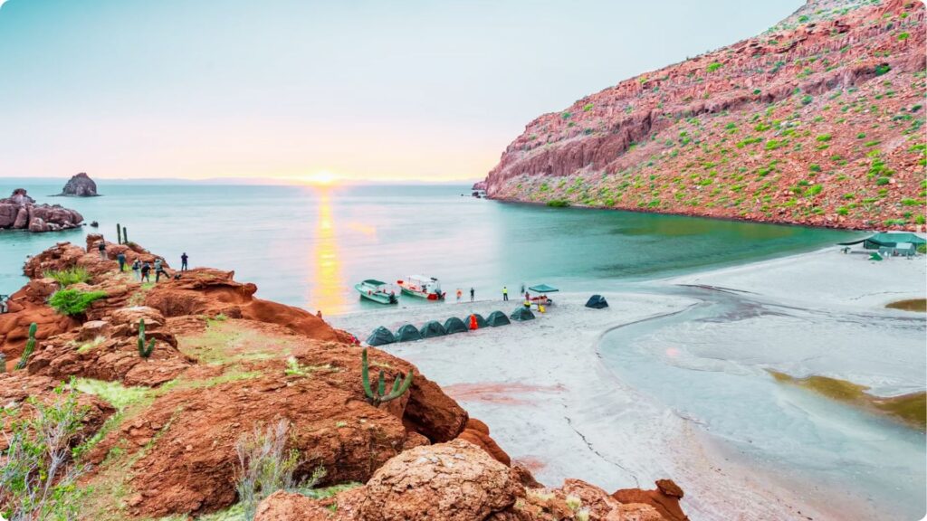 11 Best Beaches in Mexico You Need to Visit Campers and boats gather on a remote white sand beach surrounded by red desert cliffs at sunset.