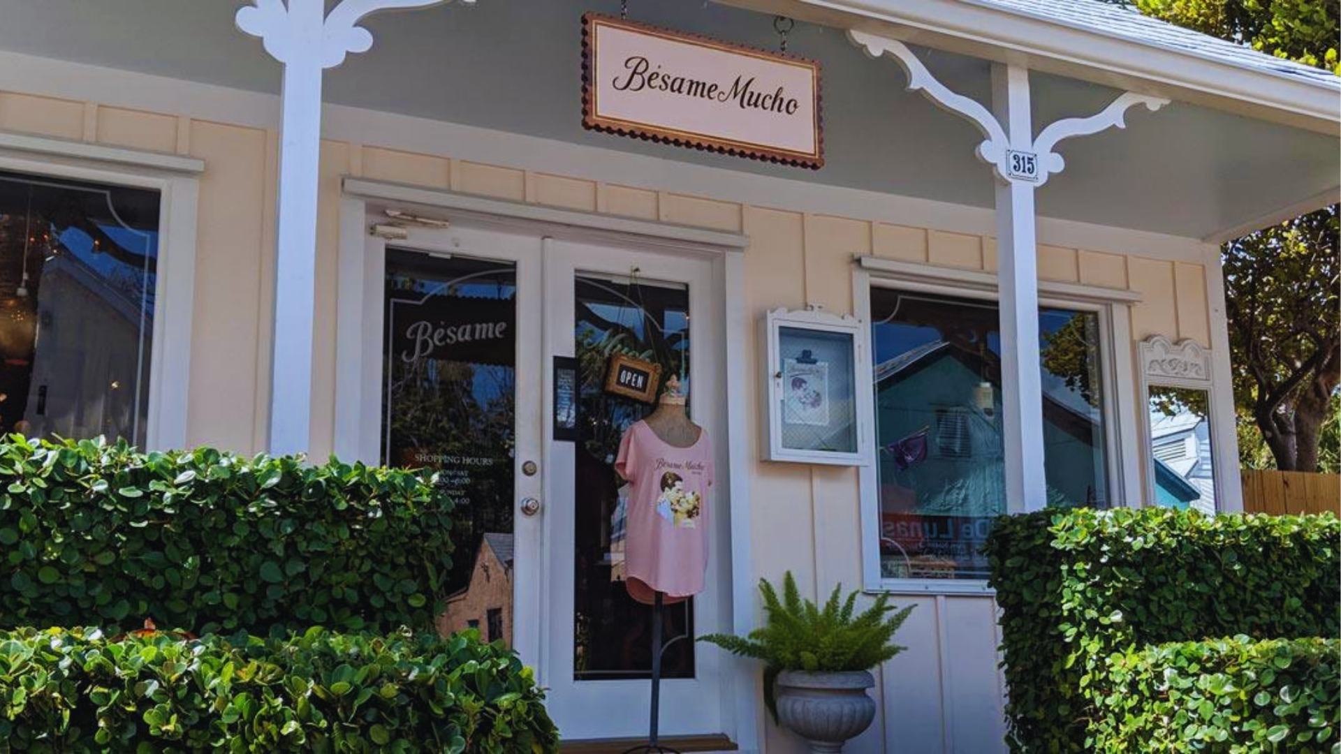 Bésame Mucho boutique storefront in Key West with Open sign, pink shirt display, white trim, and green hedges.