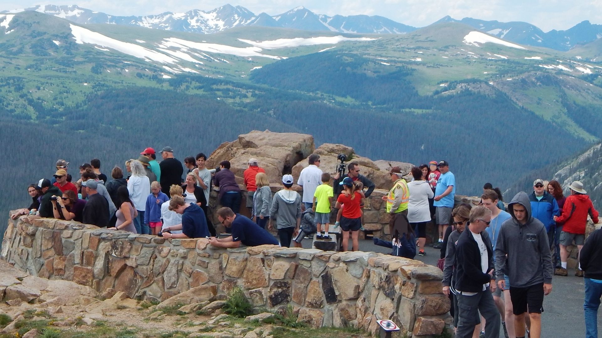 Crowds of tourists gather at mountain overlook with stone wall, snowy peaks and forested valleys in Colorado backcountry.