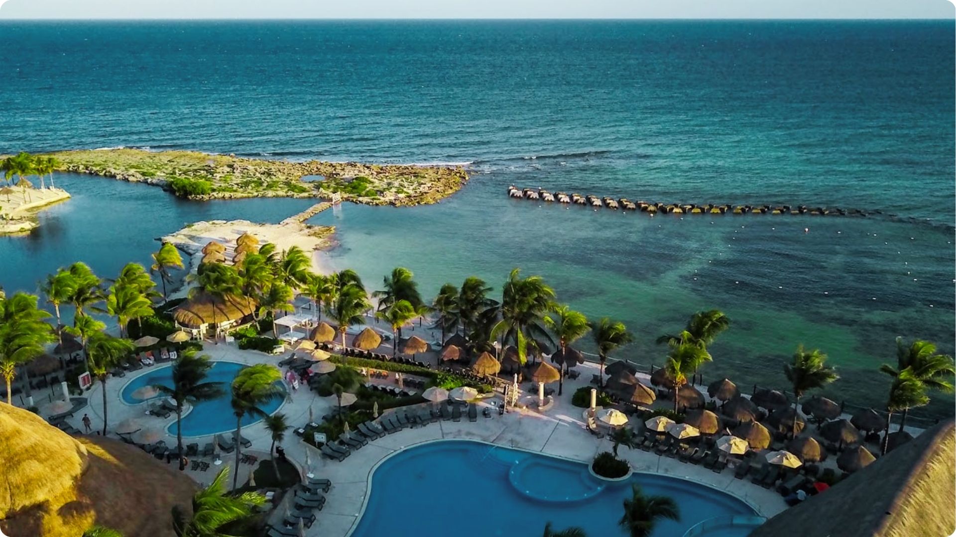 An aerial view of a luxury resort with curving pools, palm trees, and palapa umbrellas on the beach