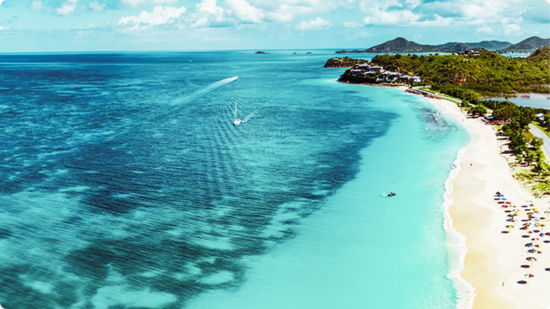 Aerial view of a long white sand beach meeting gradient turquoise water, with sailboats cruising offshore.