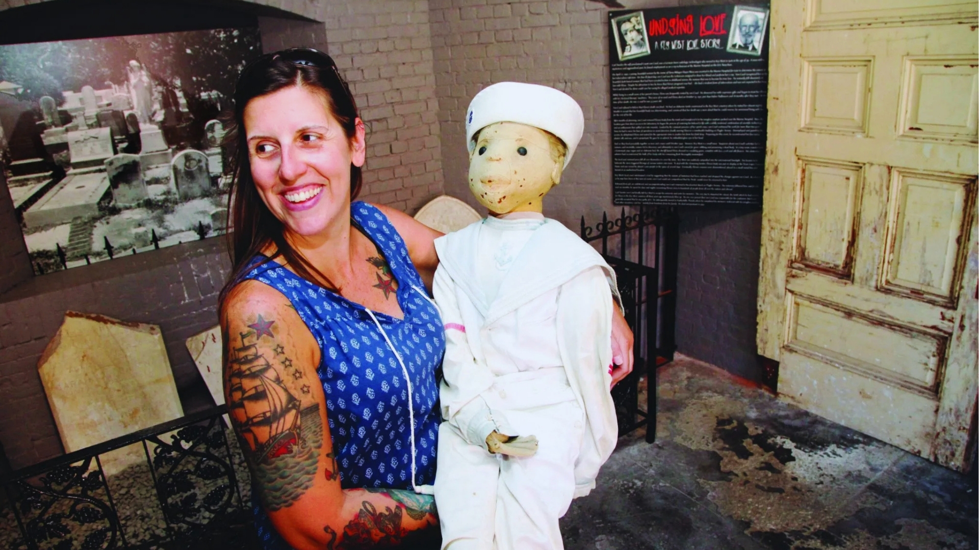 Smiling woman with tattoos holds Robert the Doll in sailor suit at Key West ghost tour museum exhibit.
