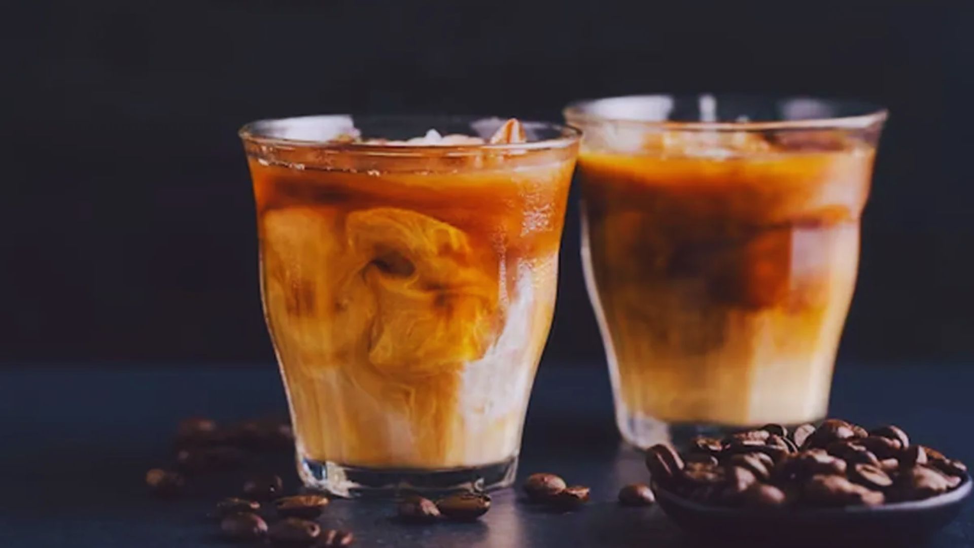 Two glasses of iced coffee sit on a table, surrounded by scattered coffee beans.