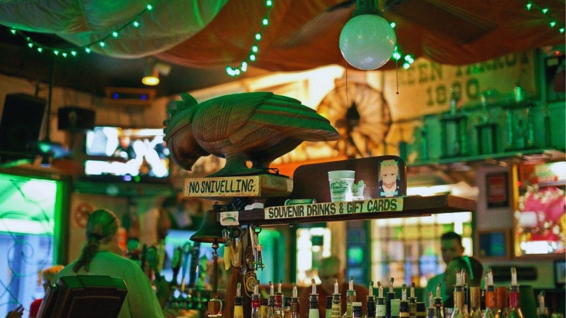 Interior of Key West Green Parrot bar with wooden rooster carving, No Snivelling sign, green lights, and liquor bottles