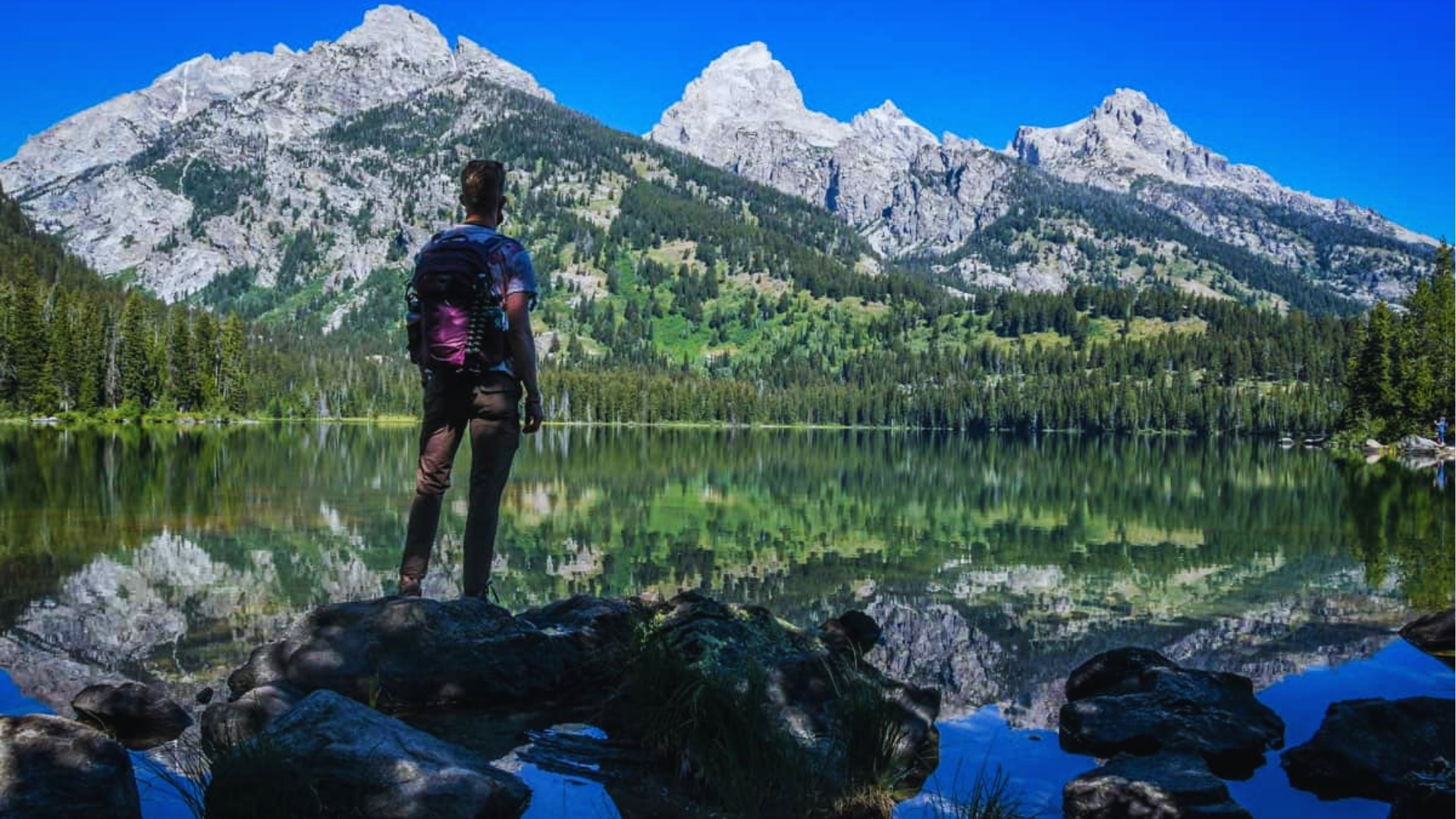 Hiker with purple backpack stands on a boulder overlooking Taggart Lake as sharp Teton peaks reflect perfectly in still water.