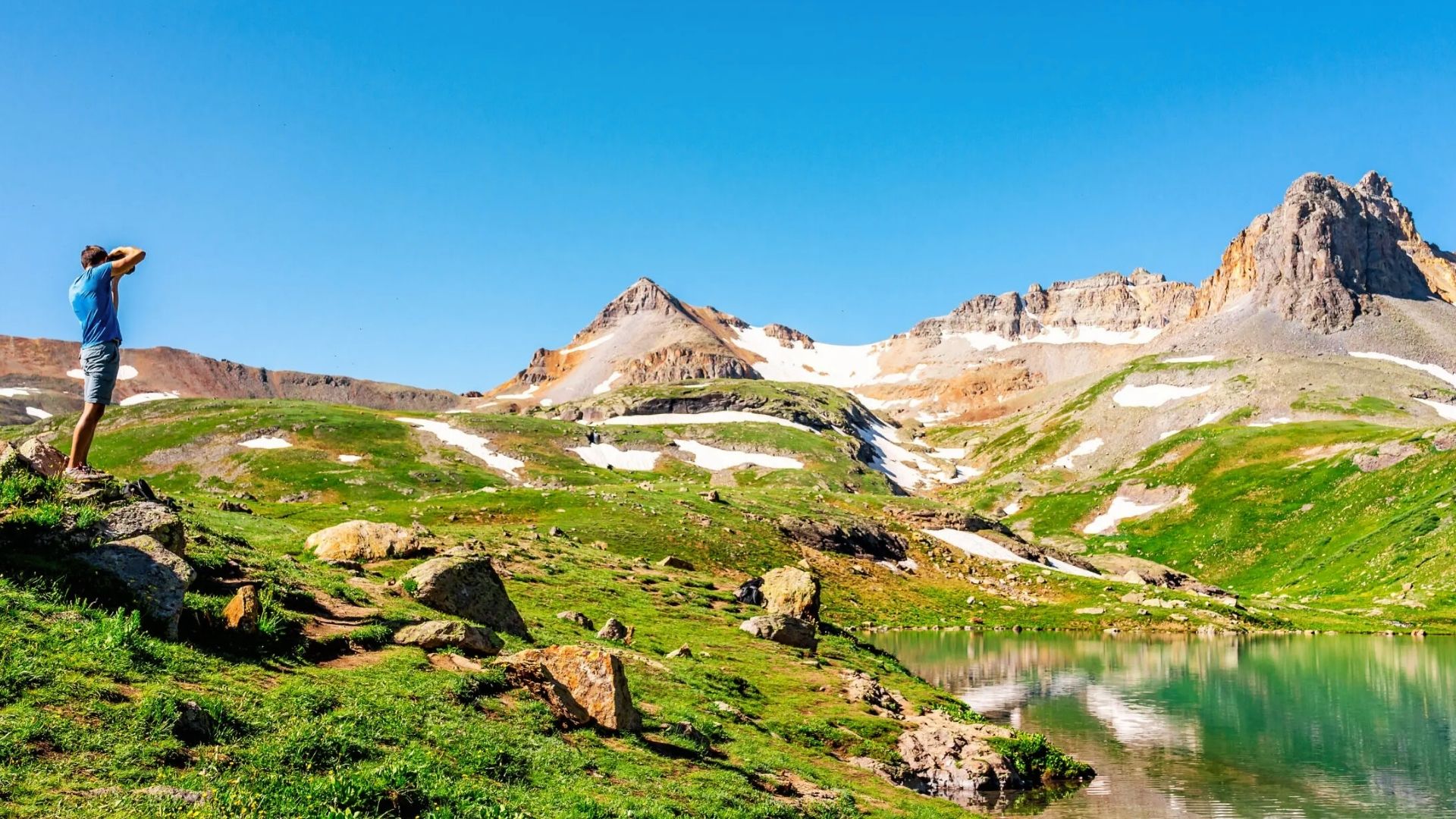 Hiker photographs turquoise Ice Lake Colorado surrounded by snow-dusted peaks and green alpine meadows under clear blue sky