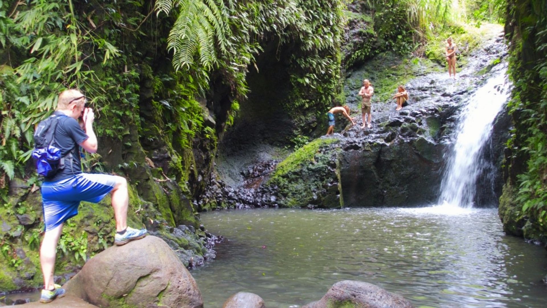 Hikers climb wet rocks and a man photographs Maunawili Falls waterfall flowing into a jungle pool
