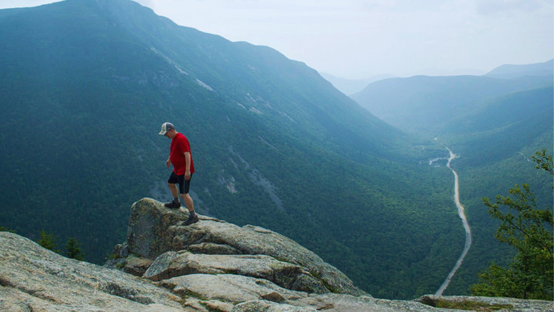 Lone hiker in red shirt stands on granite cliff edge overlooking deep green Crawford Notch valley from Mount Willard.
