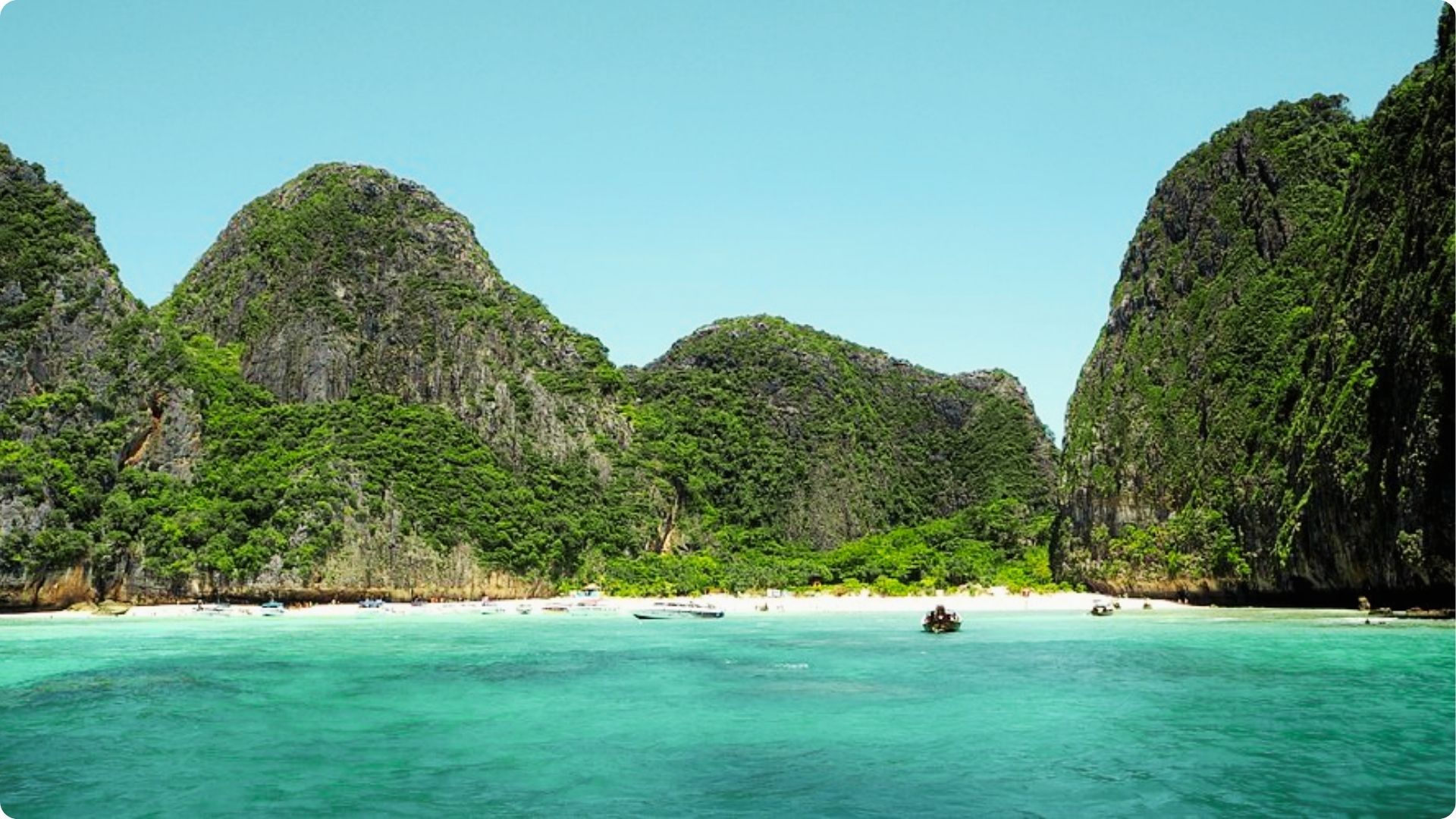 Boats float in clear turquoise water in front of a white-sand beach enclosed by towering cliffs.