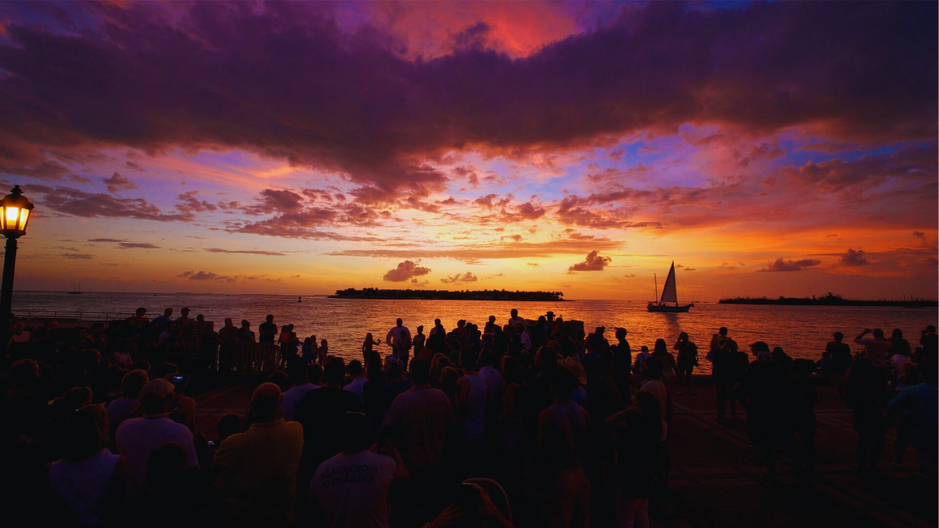 Crowd gathers at Mallory Square in Key West watching a vibrant sunset over a sailboat for a famous daily celebration.