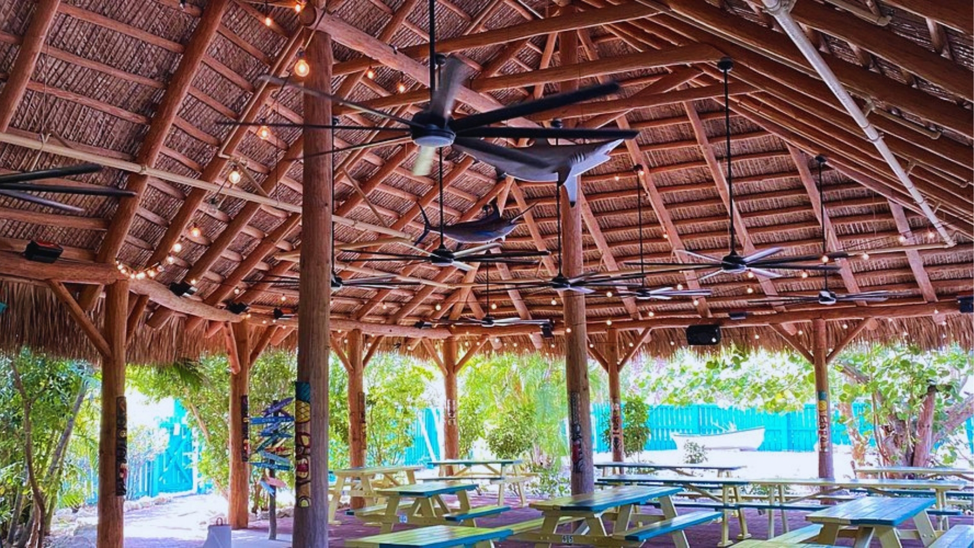 Open-air tiki pavilion with wooden beams, ceiling fans, hanging shark decor, string lights, and picnic tables in Key West.