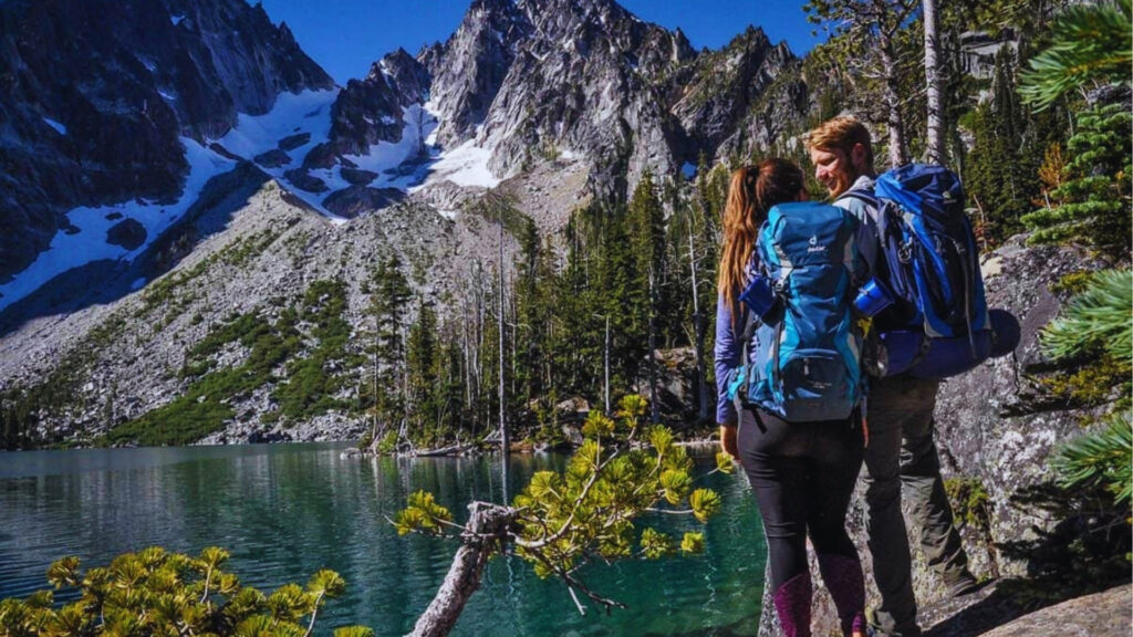 Colchuck Lake Hike Complete Trail Guide Coupled with blue backpacks stands on rocky shore of Colchuck Lake admiring steep granite peaks and glacier-carved cirque walls.