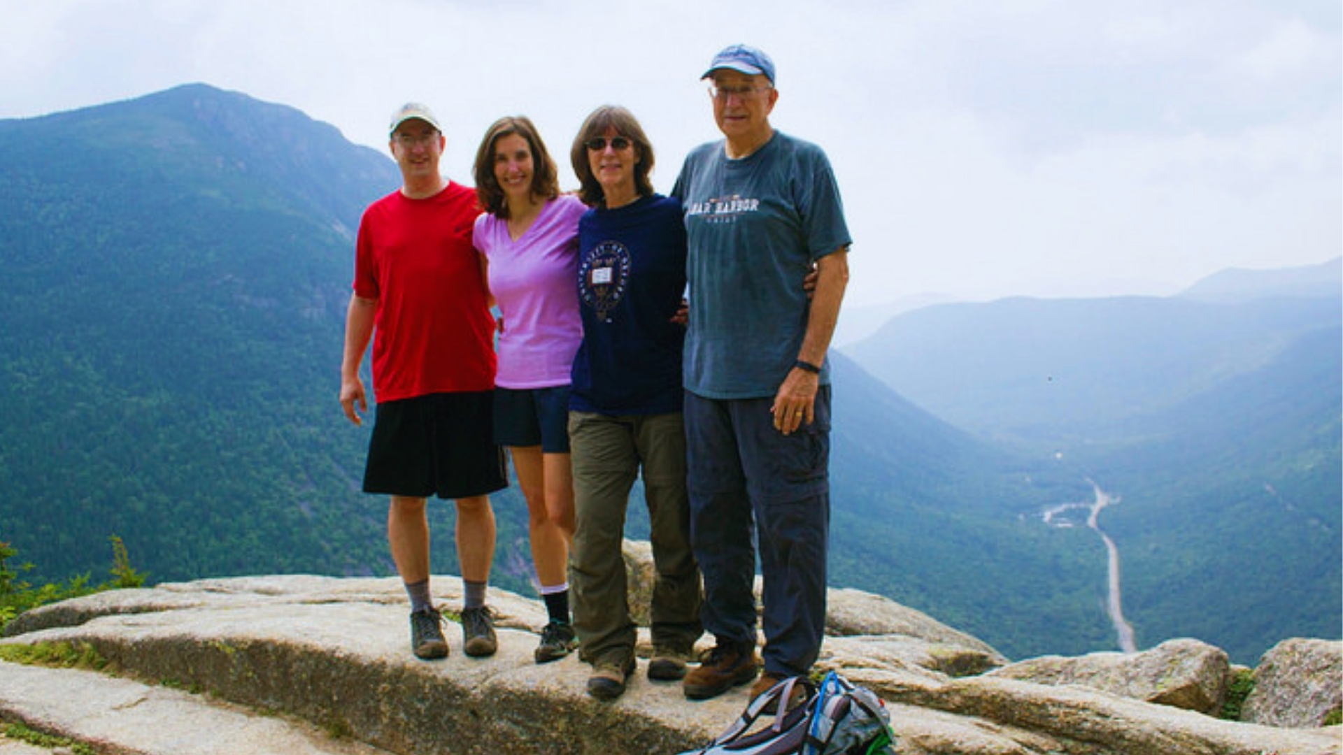 Family of four hikers poses on rocky Mount Willard summit with Crawford Notch valley, road, and forested mountains behind them.