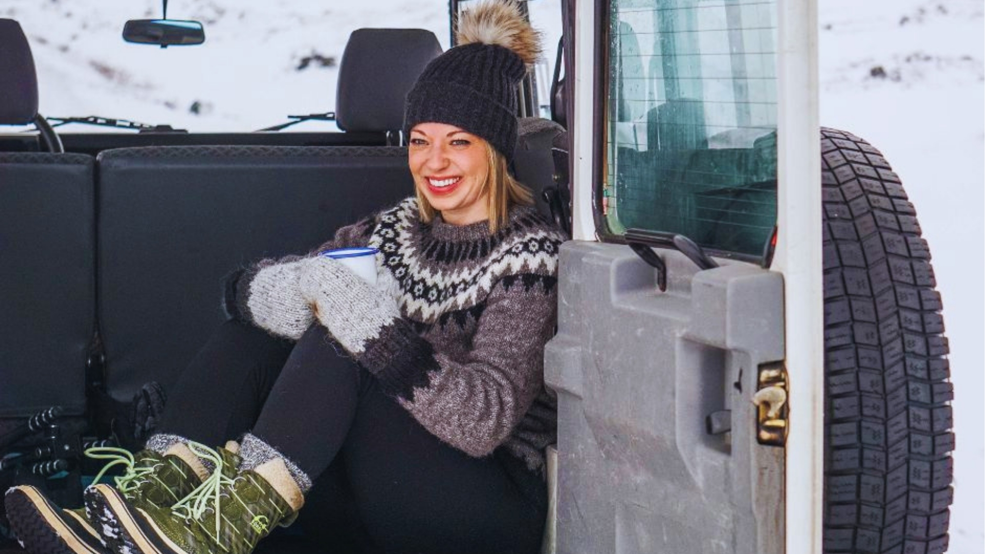 Woman in traditional Icelandic wool sweater, knit hat, and mittens smiles while holding a warm mug inside a snowy Land Rover.