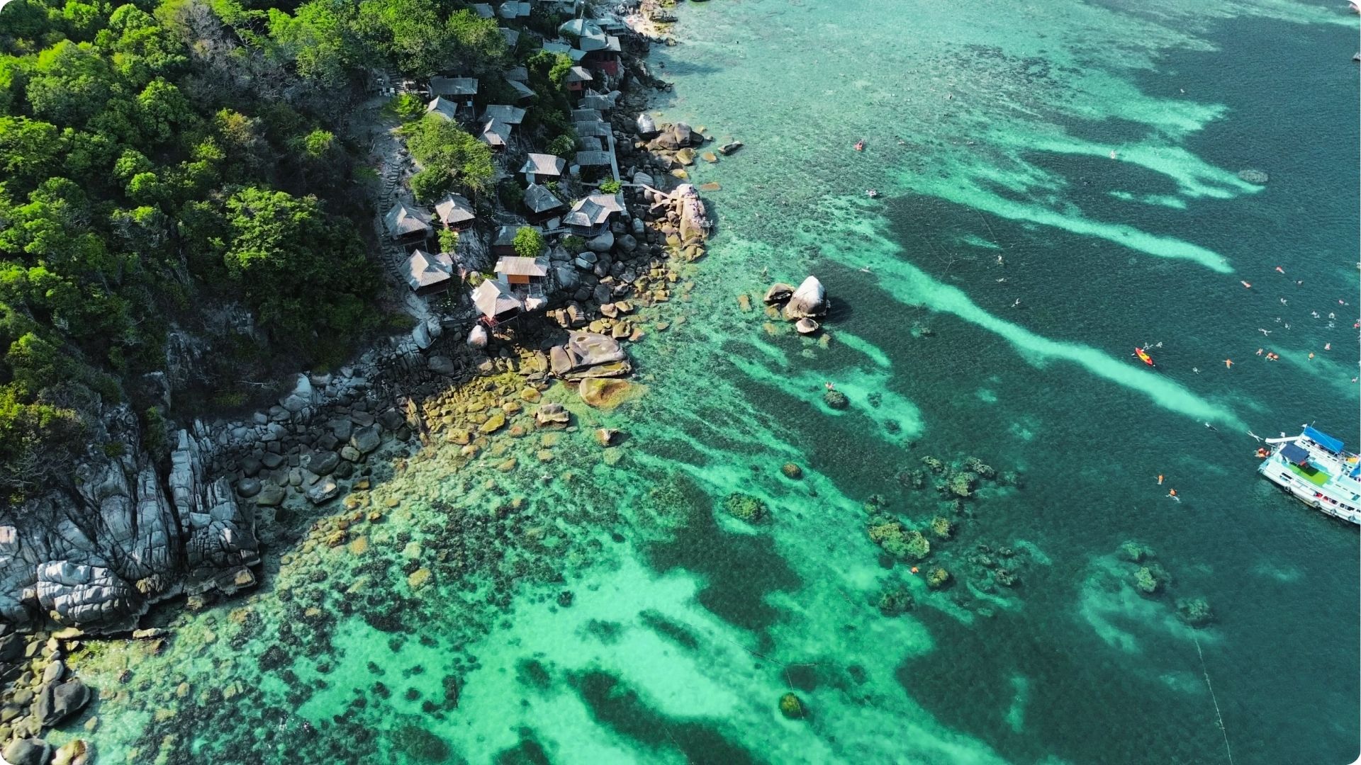 Aerial view of rustic bungalows perched on rocky cliffs above a coral-filled bay.