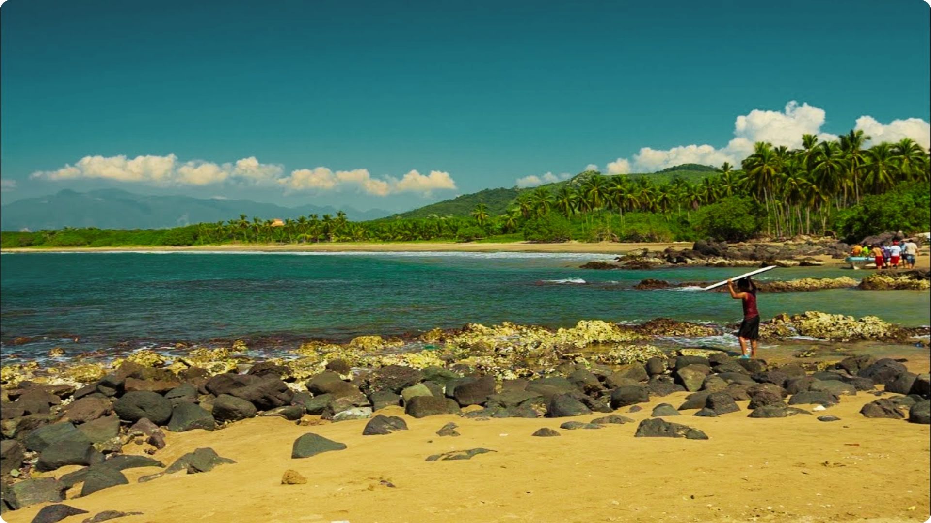 A surfer carries a board over volcanic rocks toward the calm turquoise waters of Playa Norte.