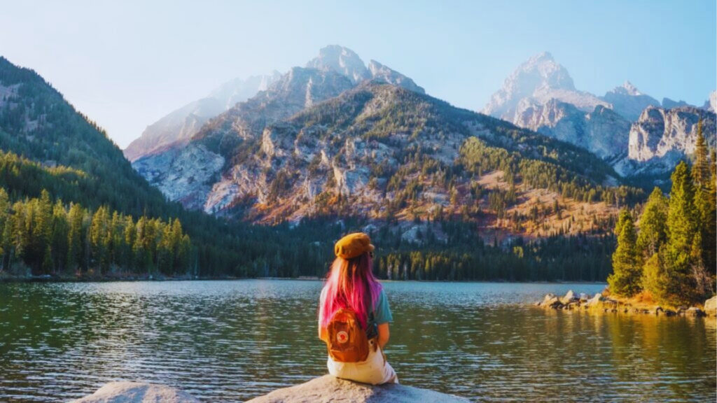 A woman with pink hair and brown hat sits on a rock facing Taggart Lake at sunset with Tetons glowing gold.