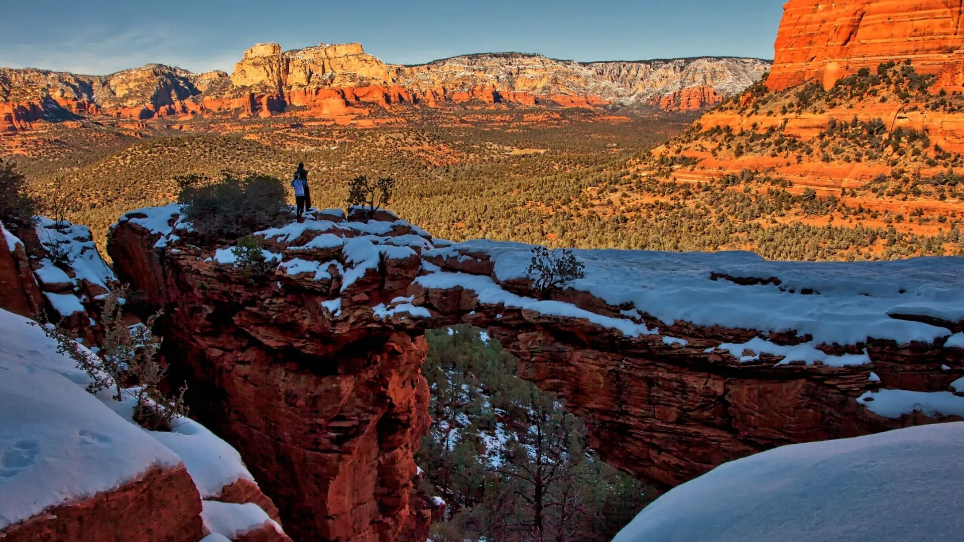 Snow covered Devils Bridge arch in winter with two hikers and panoramic Sedona red rock mountains