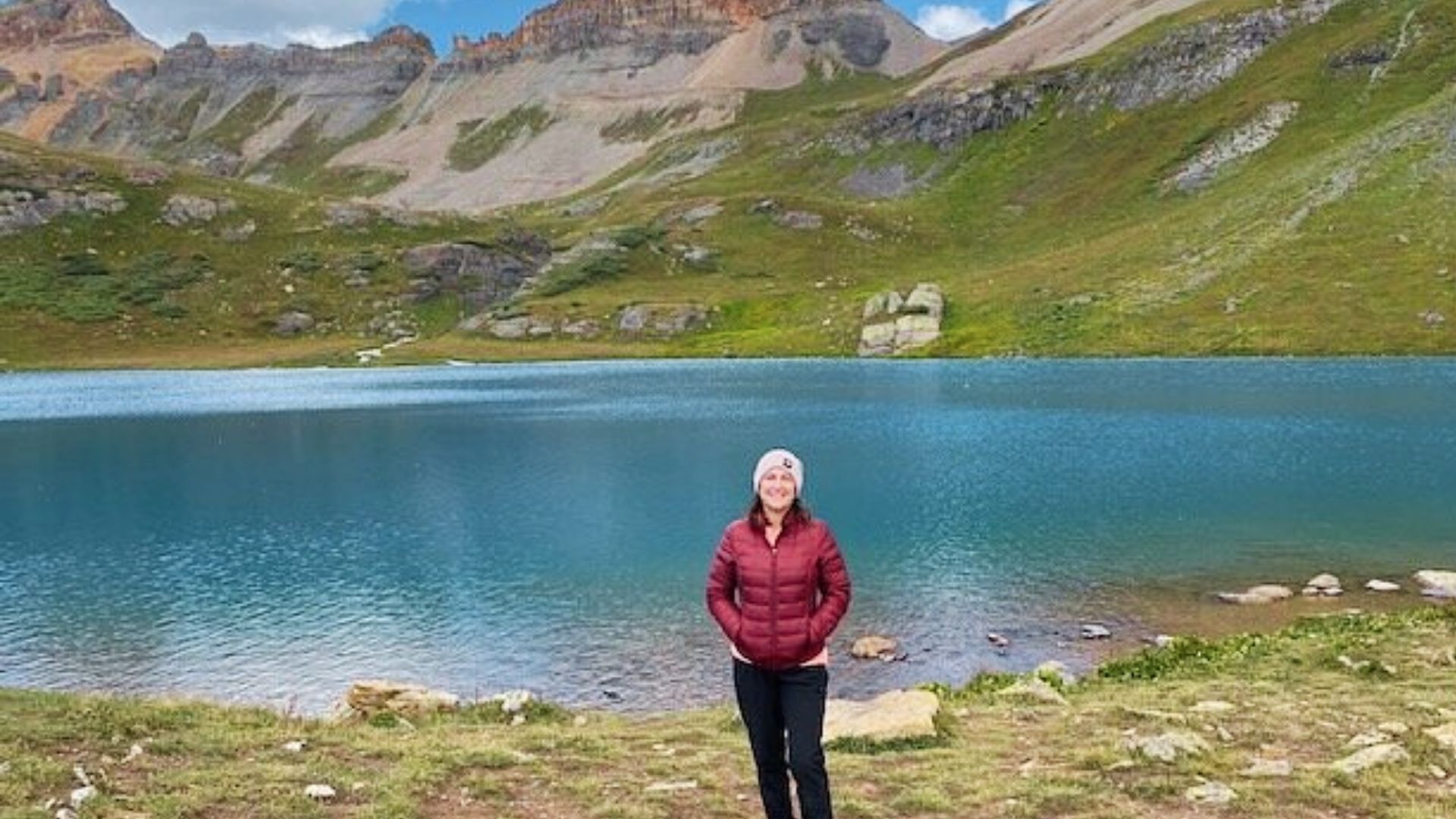 Woman in red jacket and beanie smiles at Ice Lake Colorado shoreline with vibrant blue water and rugged mountain backdrop