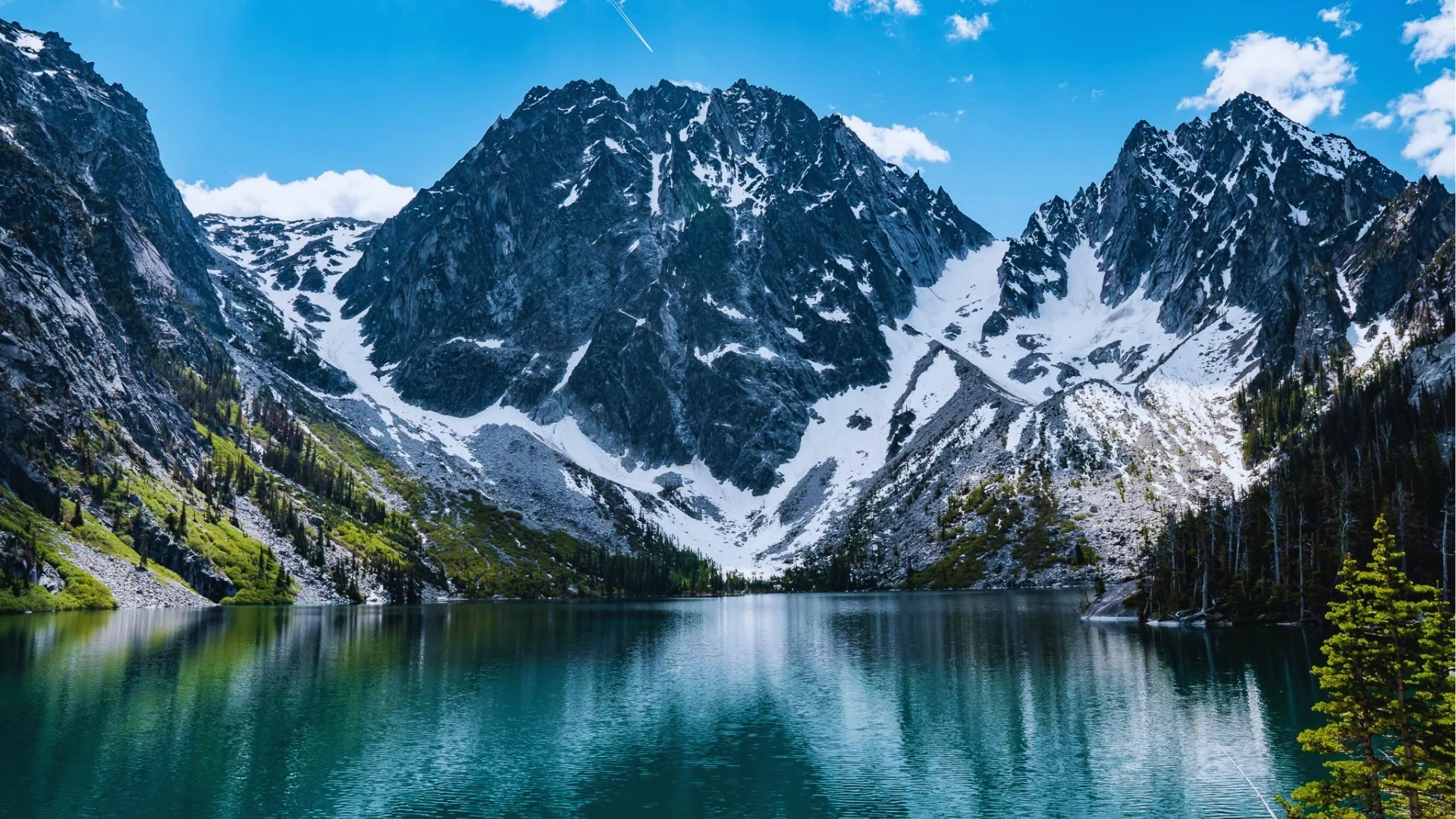 Pristine turquoise Colchuck Lake reflects jagged snow-covered Dragontail and Colchuck Peaks under blue sky with pine trees lining shore